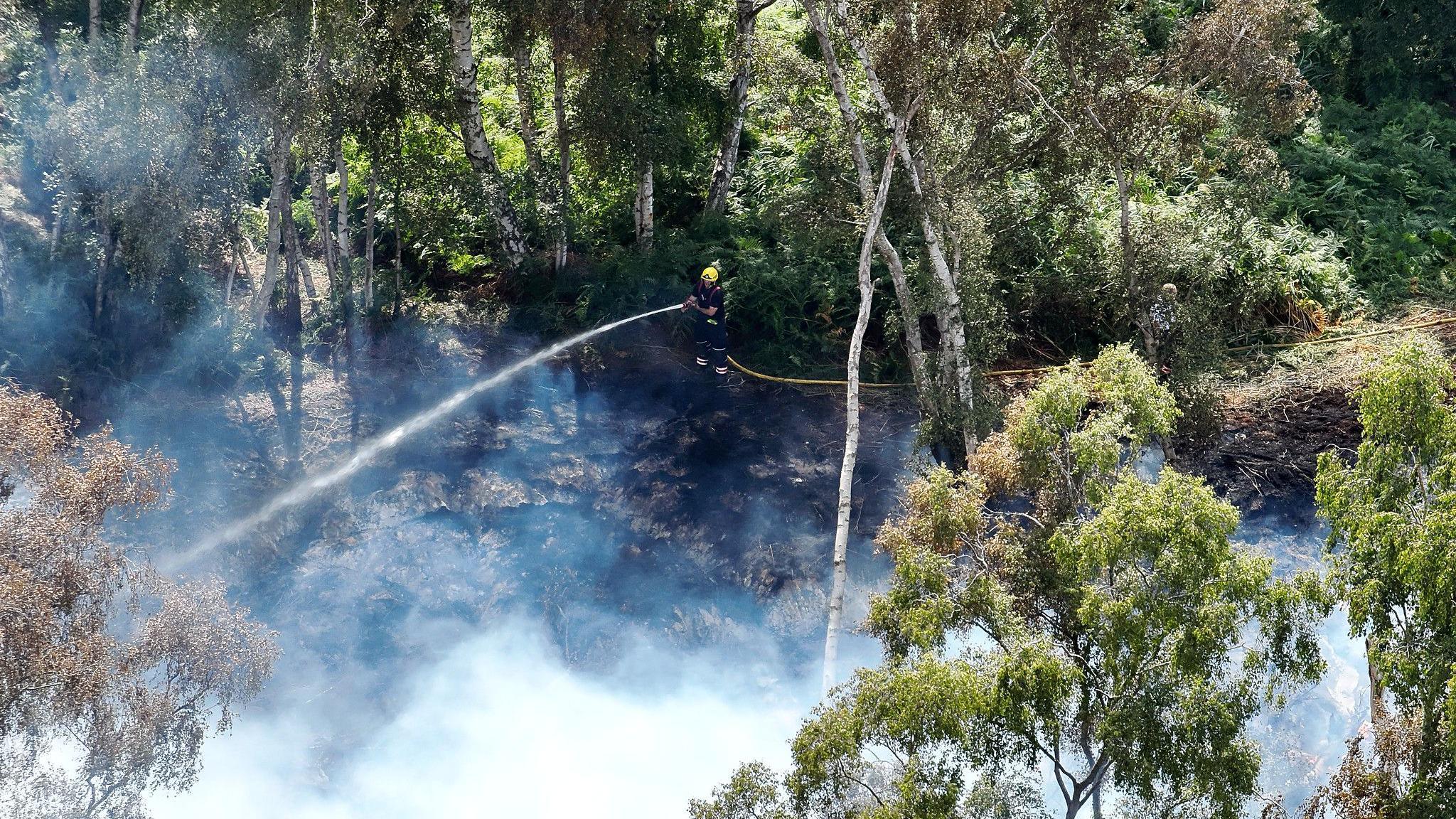 A large green space on fire with grey plumes of smoke and a firefighter in a blue uniform and yellow helmet using a hosepipe to put the fire out.