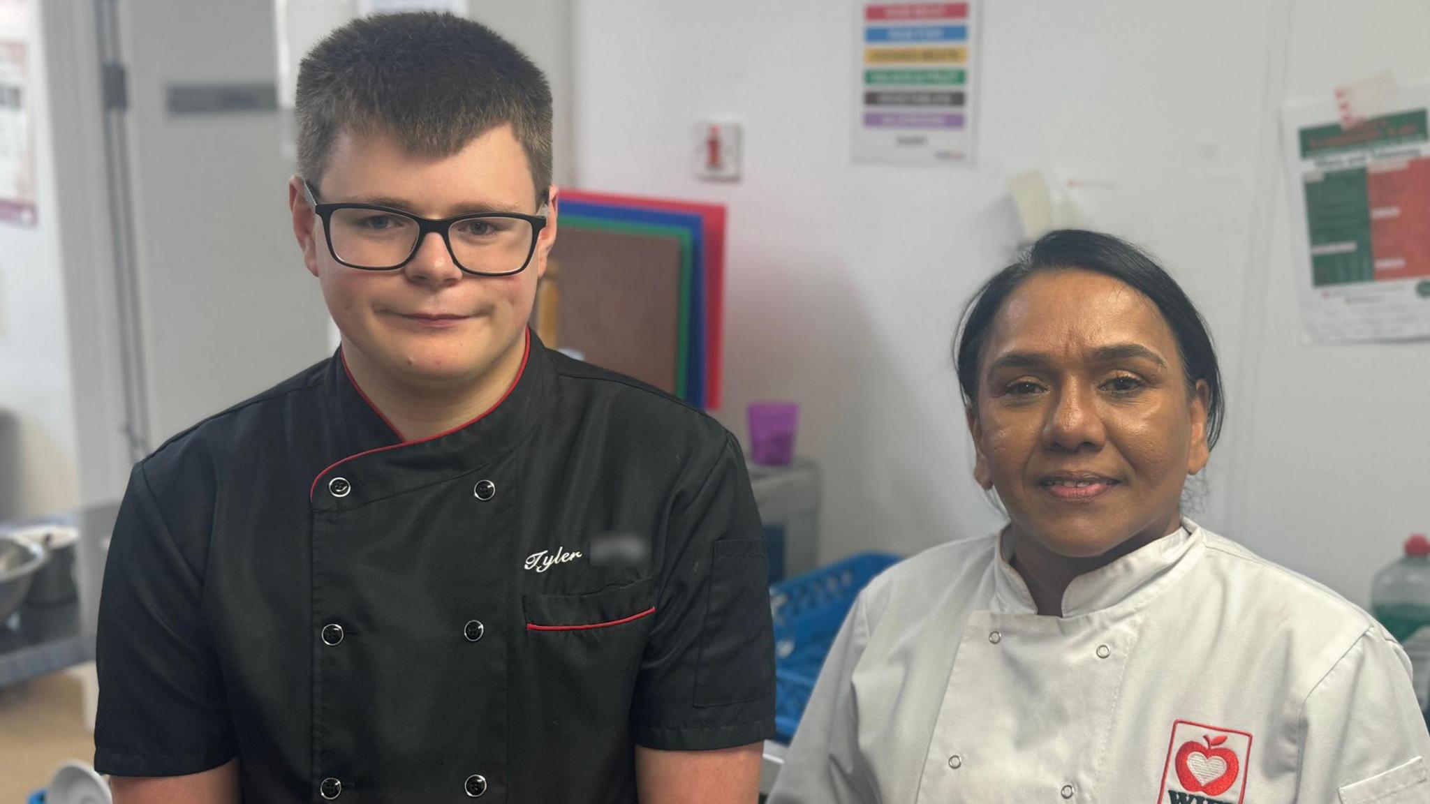 Tyler, left, has short dark hair and is wearing dark rimmed glasses and a black chef's uniform. Anna has dark hair pulled back in a pony tail and is wearing a white chef's uniform.