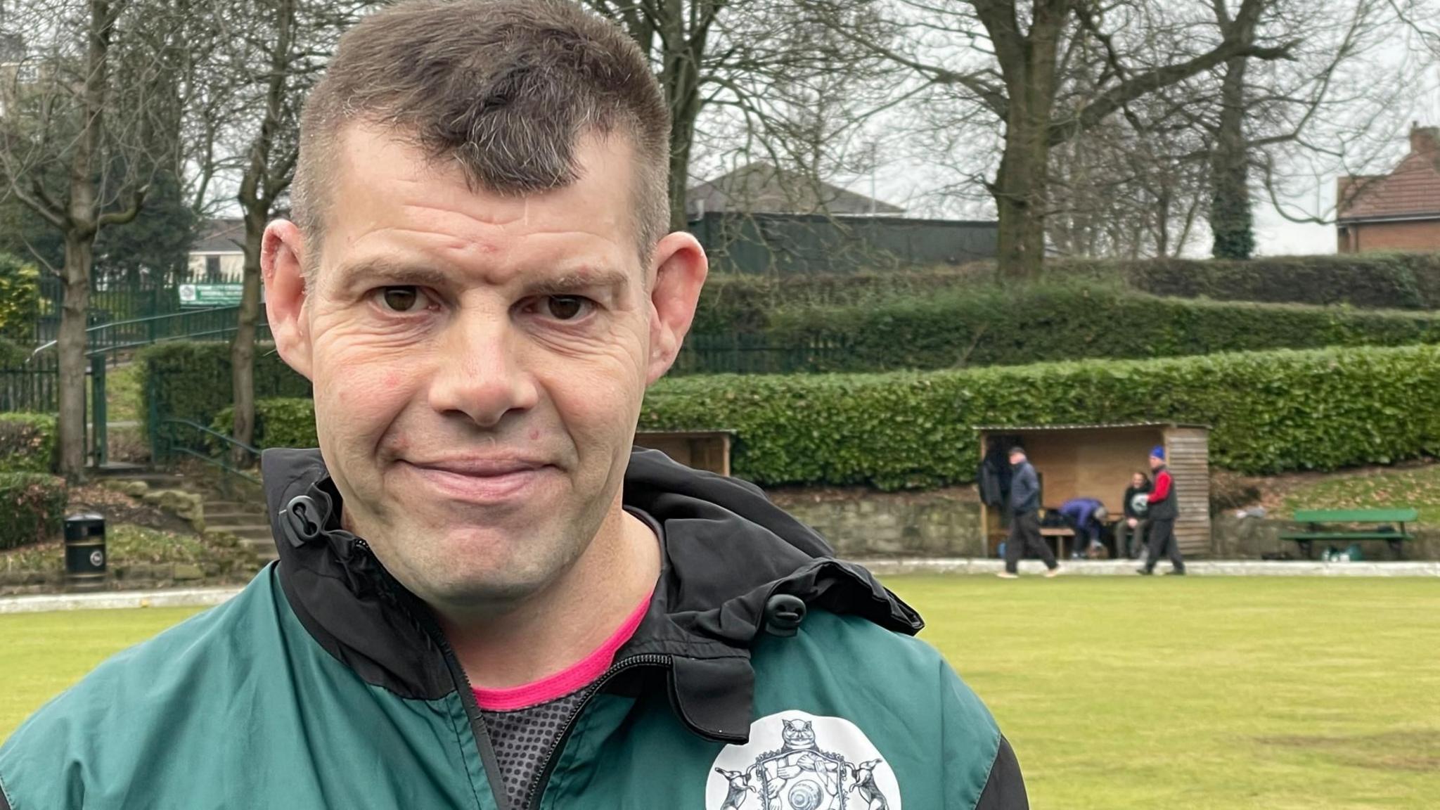 A man with short brown hair wearing a pink and grey top and a green and black waterproof jacket standing on a bowling green.