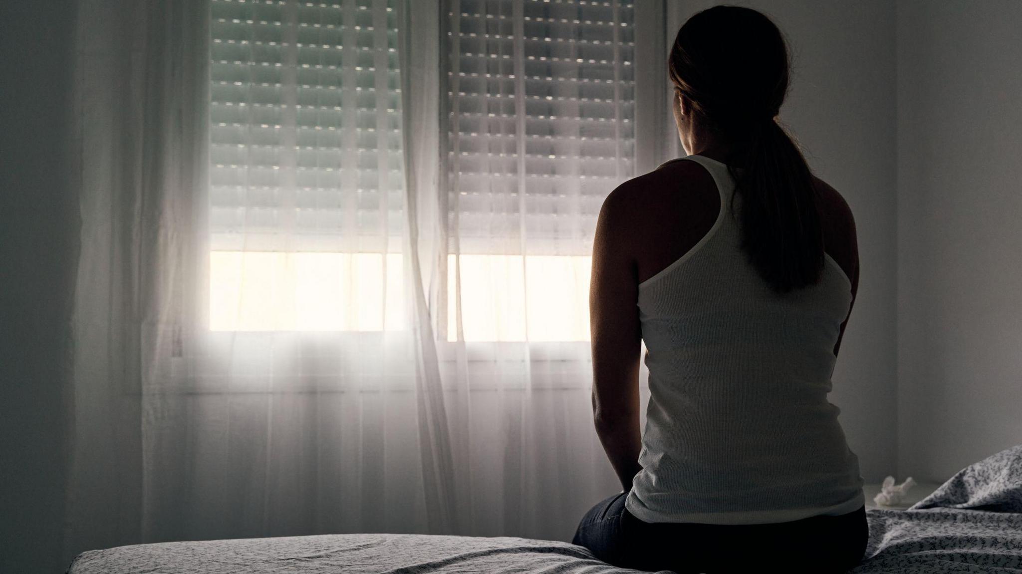 A woman has her back to the camera and is sitting on the edge of a bed in a dark room, facing the window. She has her hair tied back into a ponytail and is wearing jeans and a white tank top.