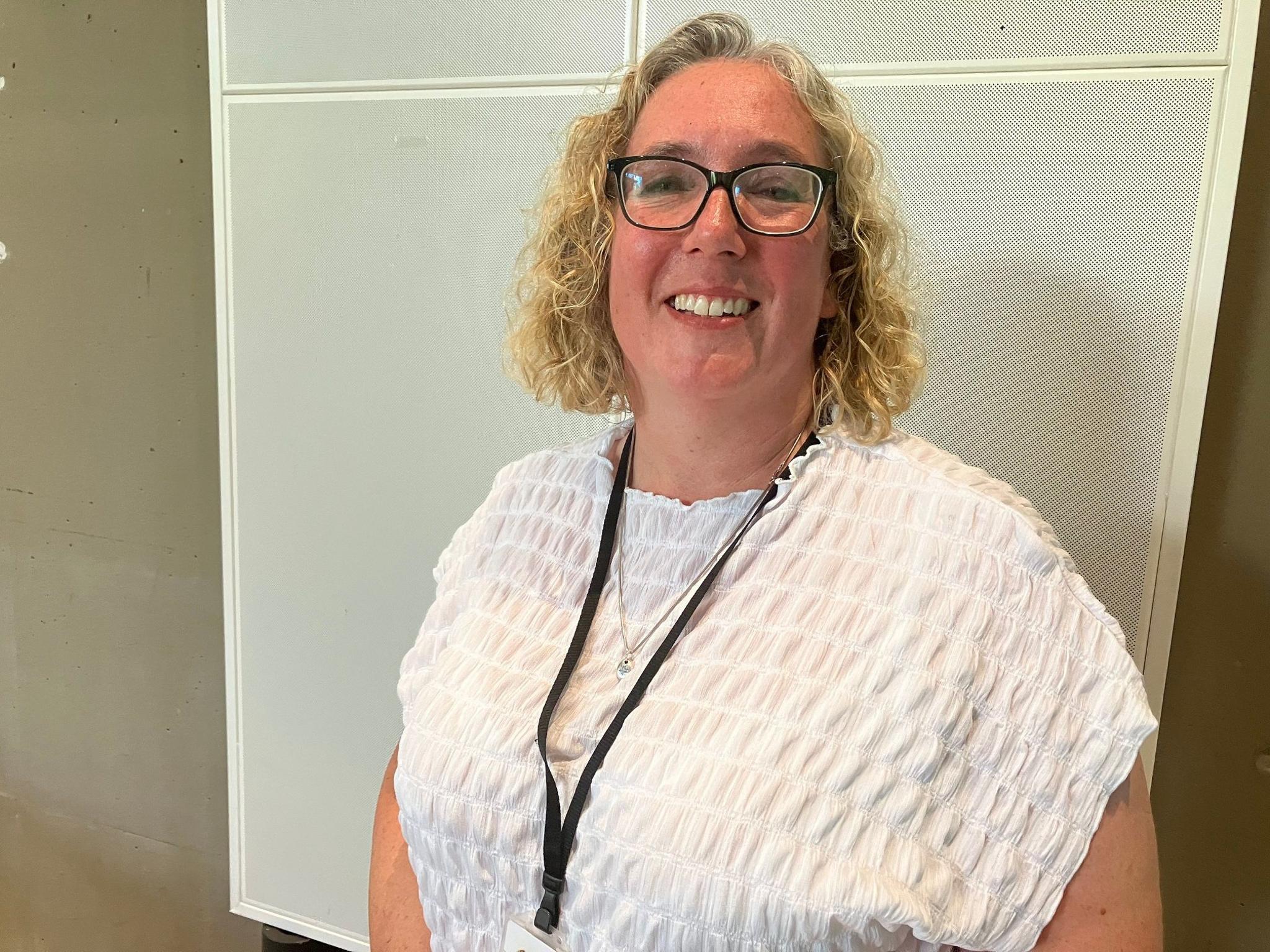 Hayley Whitaker, who has curly blonde hair, glasses and is wearing a white top, smiles at the camera. She is wearing a lanyard.