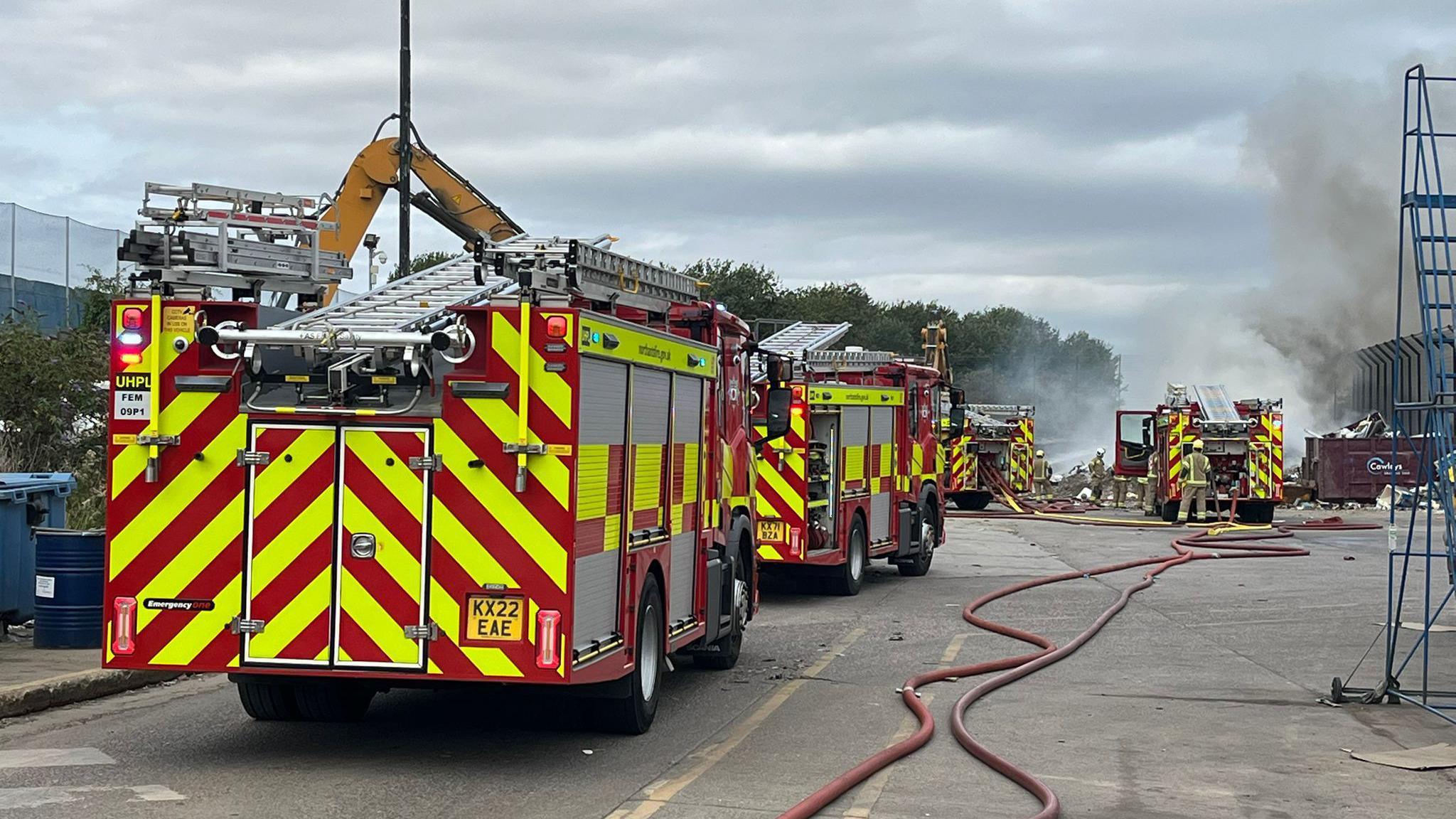 Four fire engines are parked behind a plume of grey and white smoke. Two red hoses are along the floor going towards the direction of the smoke.