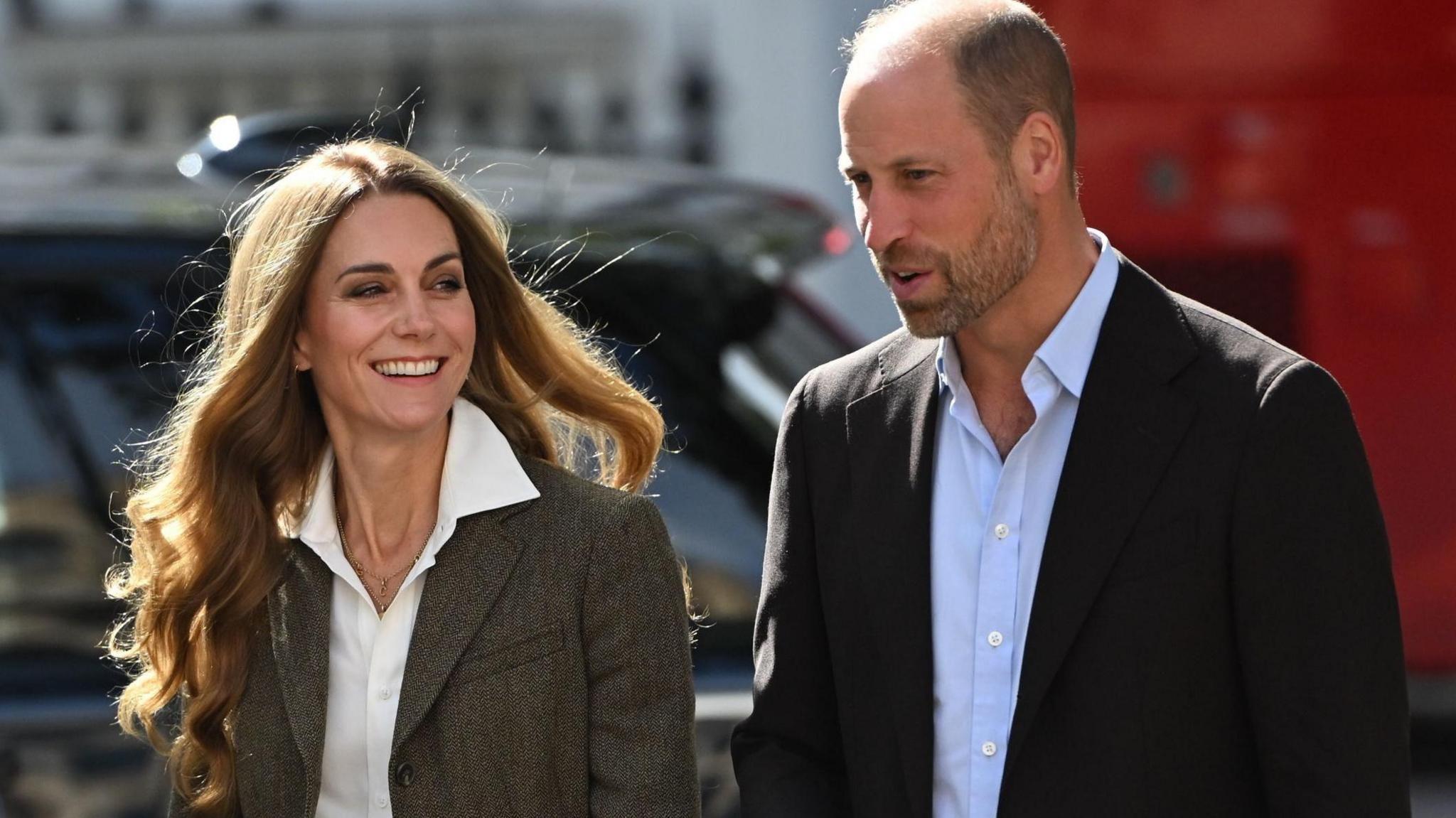 Shoulder crop of William and Catherine. William is wearing a black blazer and light blue shirt. Catherine is wearing a tweed-style jacket and white shirt. She has blonde hair. Both are smiling and posing outside the museum.