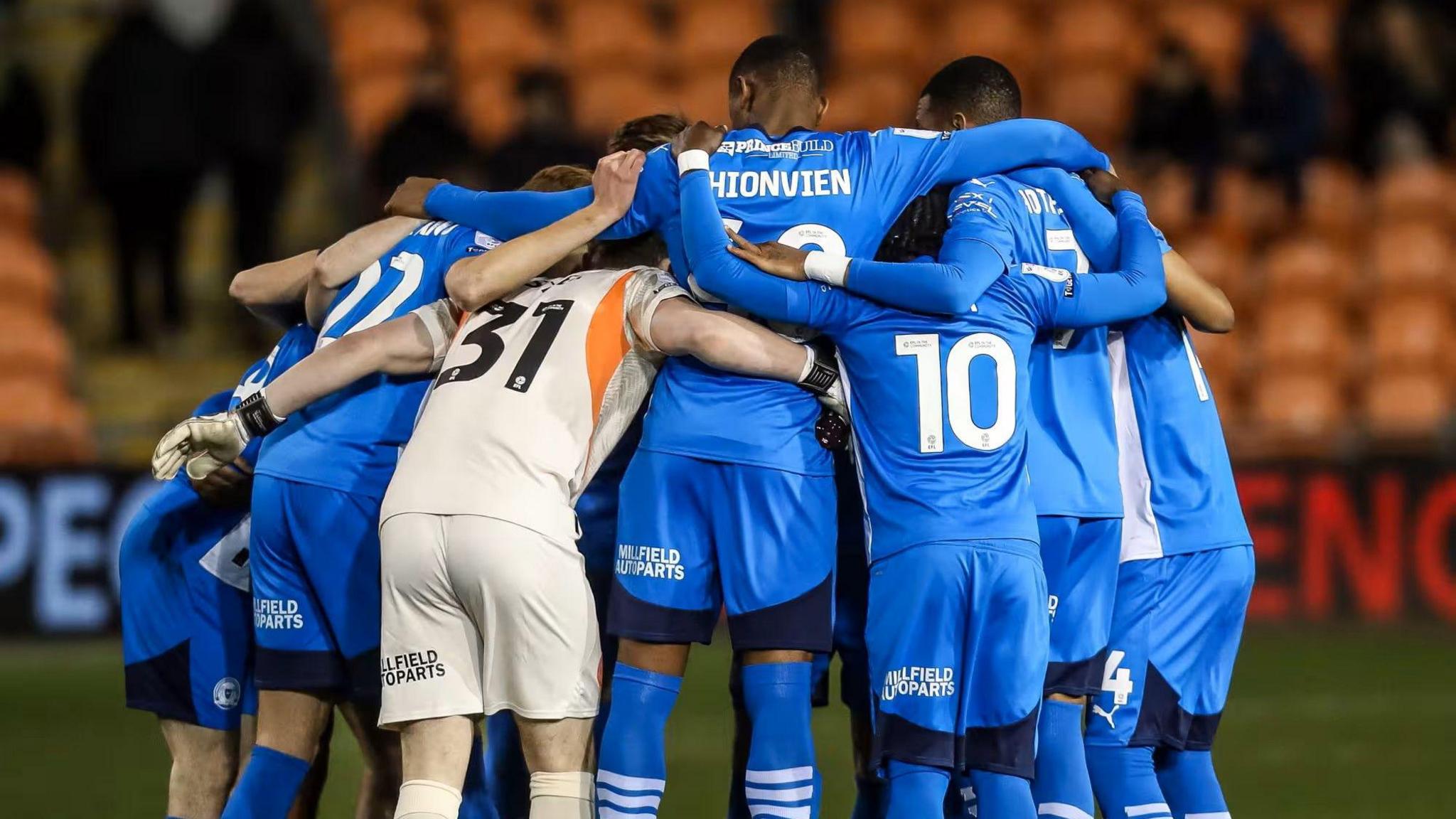 The Peterborough United team face away from the camera in a huddle with their arms across each other's shoulders.