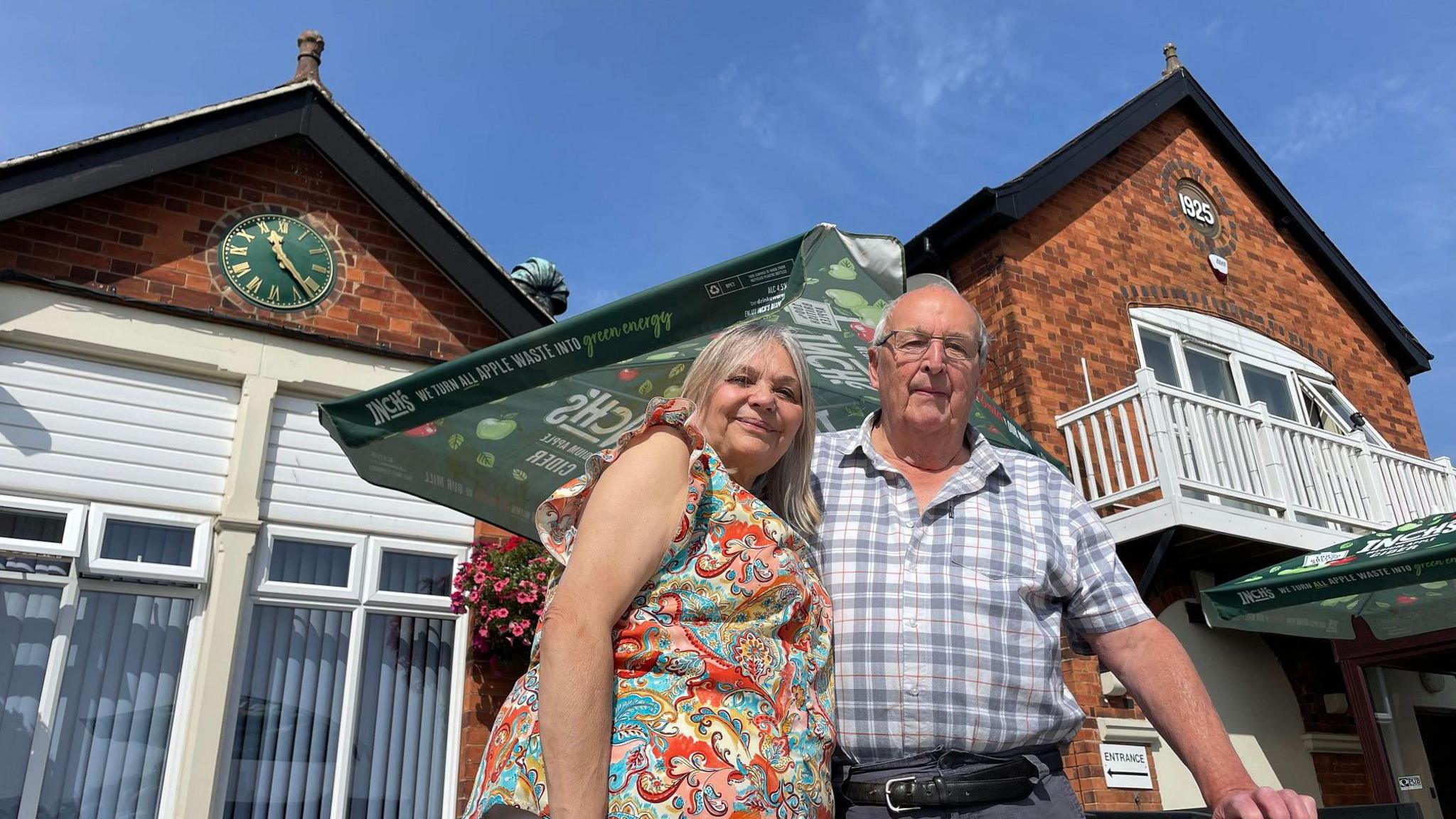 Susan and Alan outside on the terrace overlooking the bowling green, with the building behind them