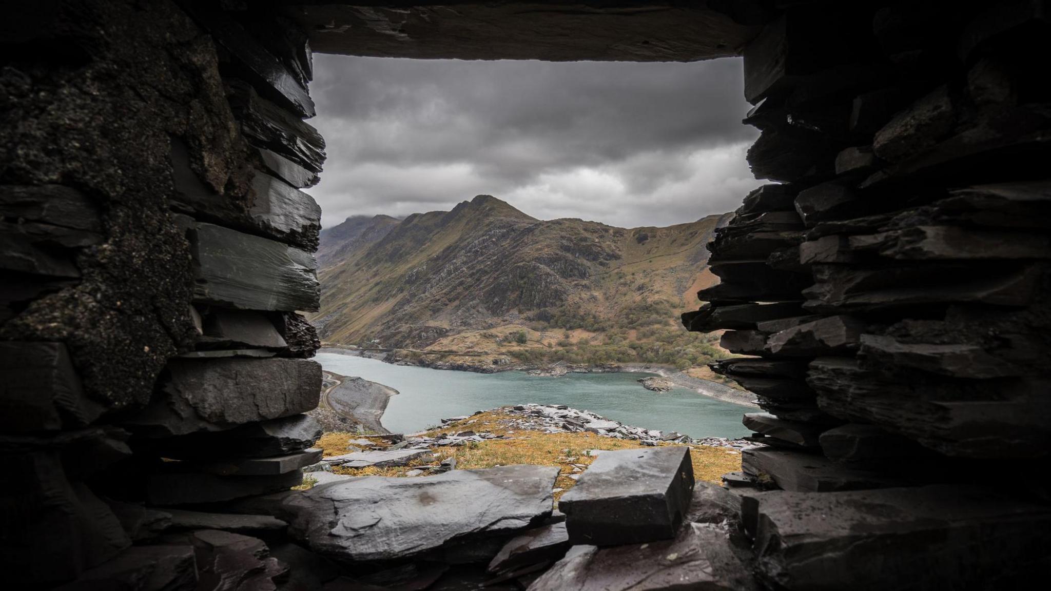 Looking through a grey glassless window in a slate building. It frames a view of mountains in the Eryri range, looking over the blue Llyn Peris lake, with a peak rising in the back, The sky is dark with heavy black clouds, giving the whole image a foreboding feel. It looks bleak and dangerous.