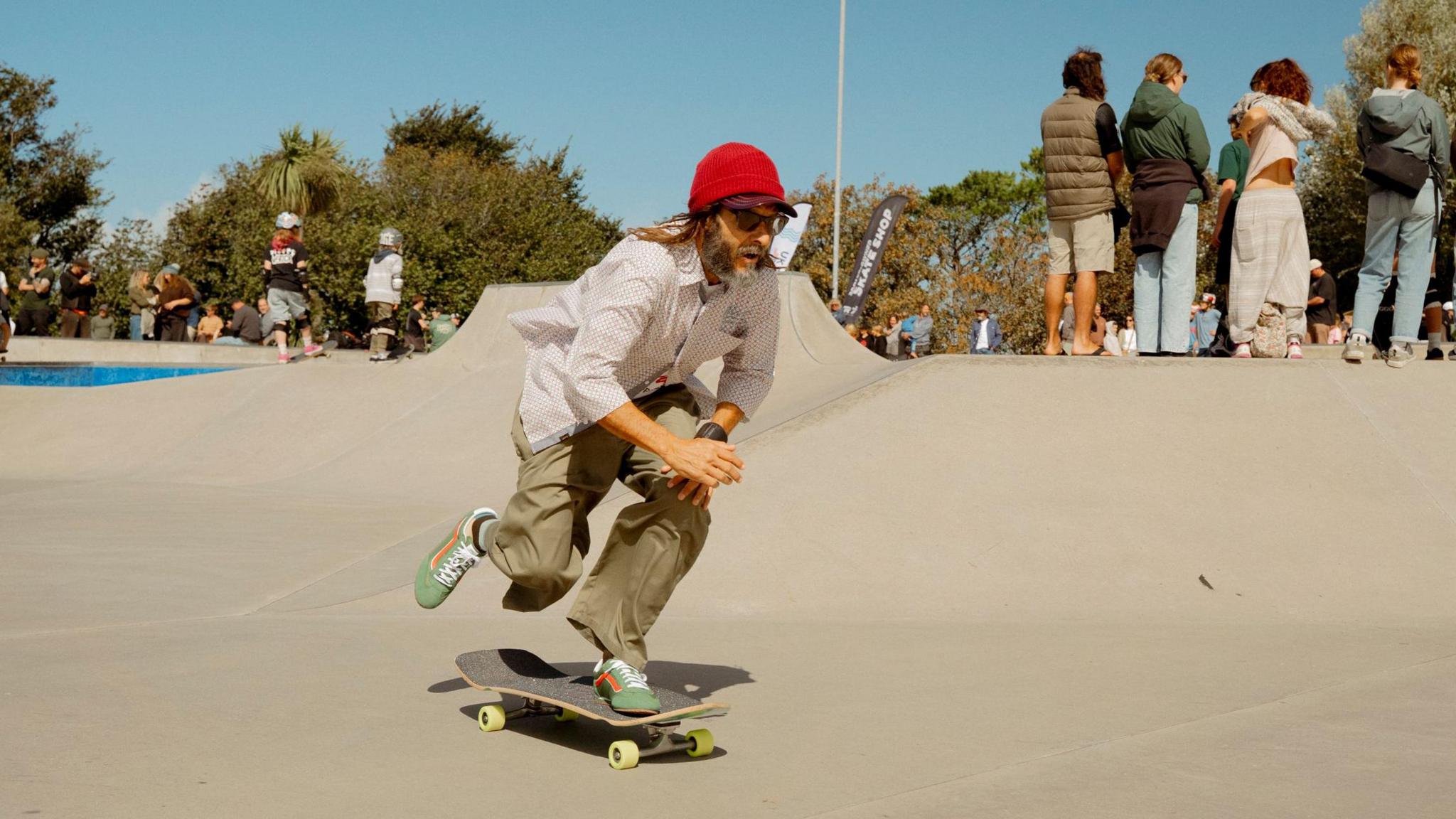 This picture shows a person skateboarding in a skate park. They're wearing a red beanie, white long-sleeve shirt, khaki trousers, and green trainers. The skateboarder is actively pushing off the ground with one foot while the other remains on the board. In the background, several people are standing or sitting.
