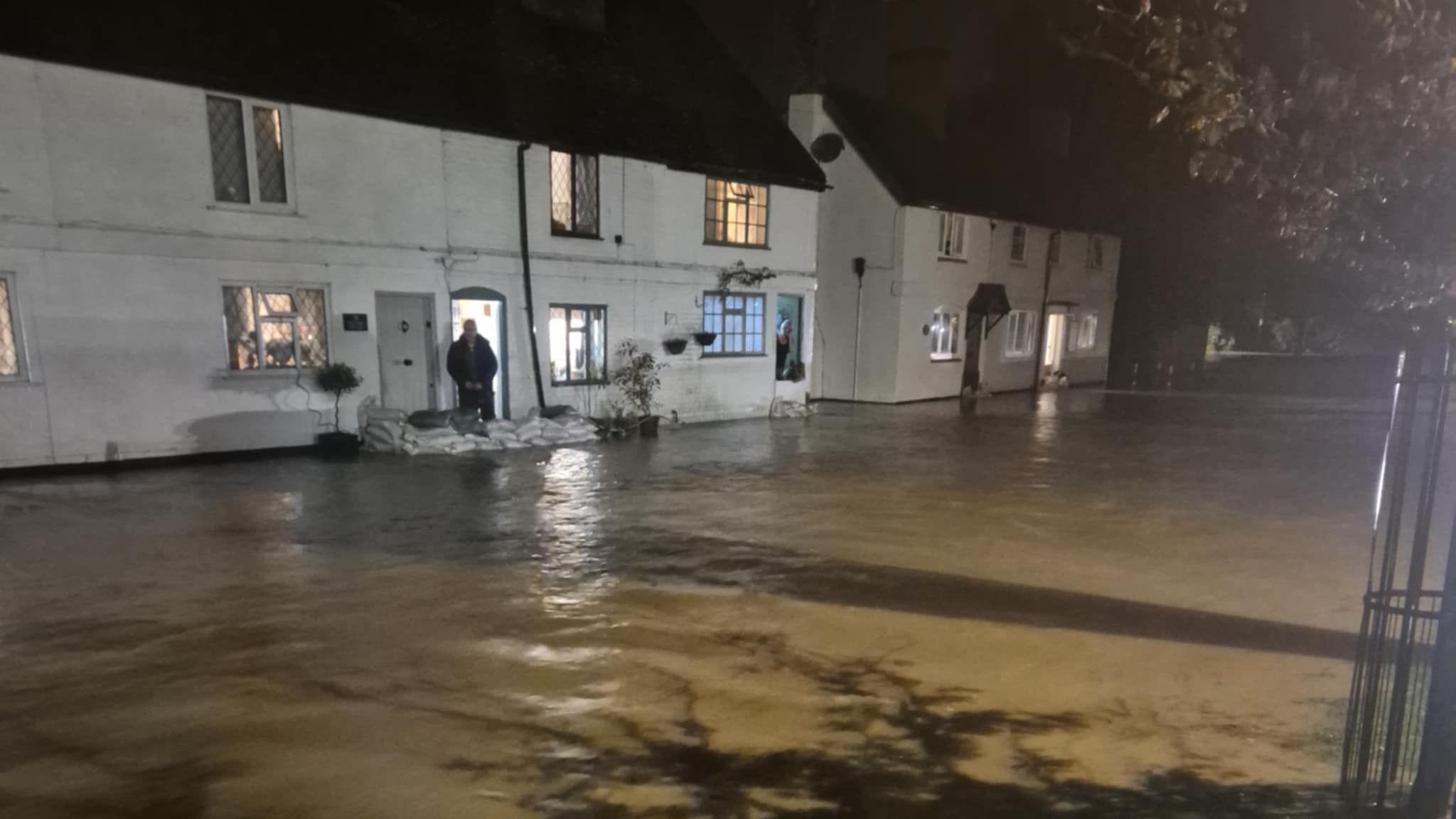 A row of white houses, one with sandbags outside the front with brown flood water across the road