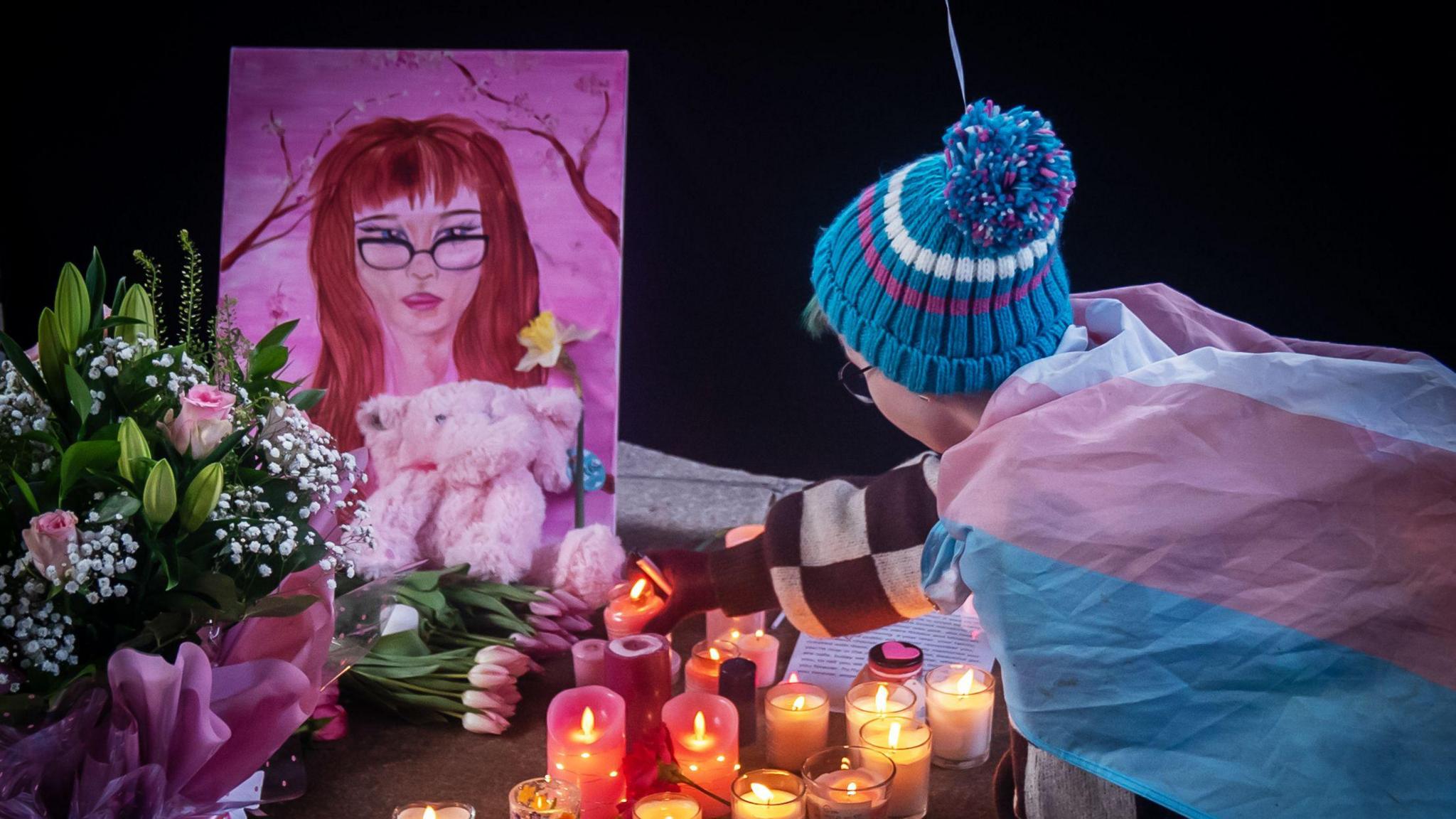 A child leaves a candle at a tribute for Brianna. Flowers are on the ground, with a pink teddy and pink drawing of Brianna