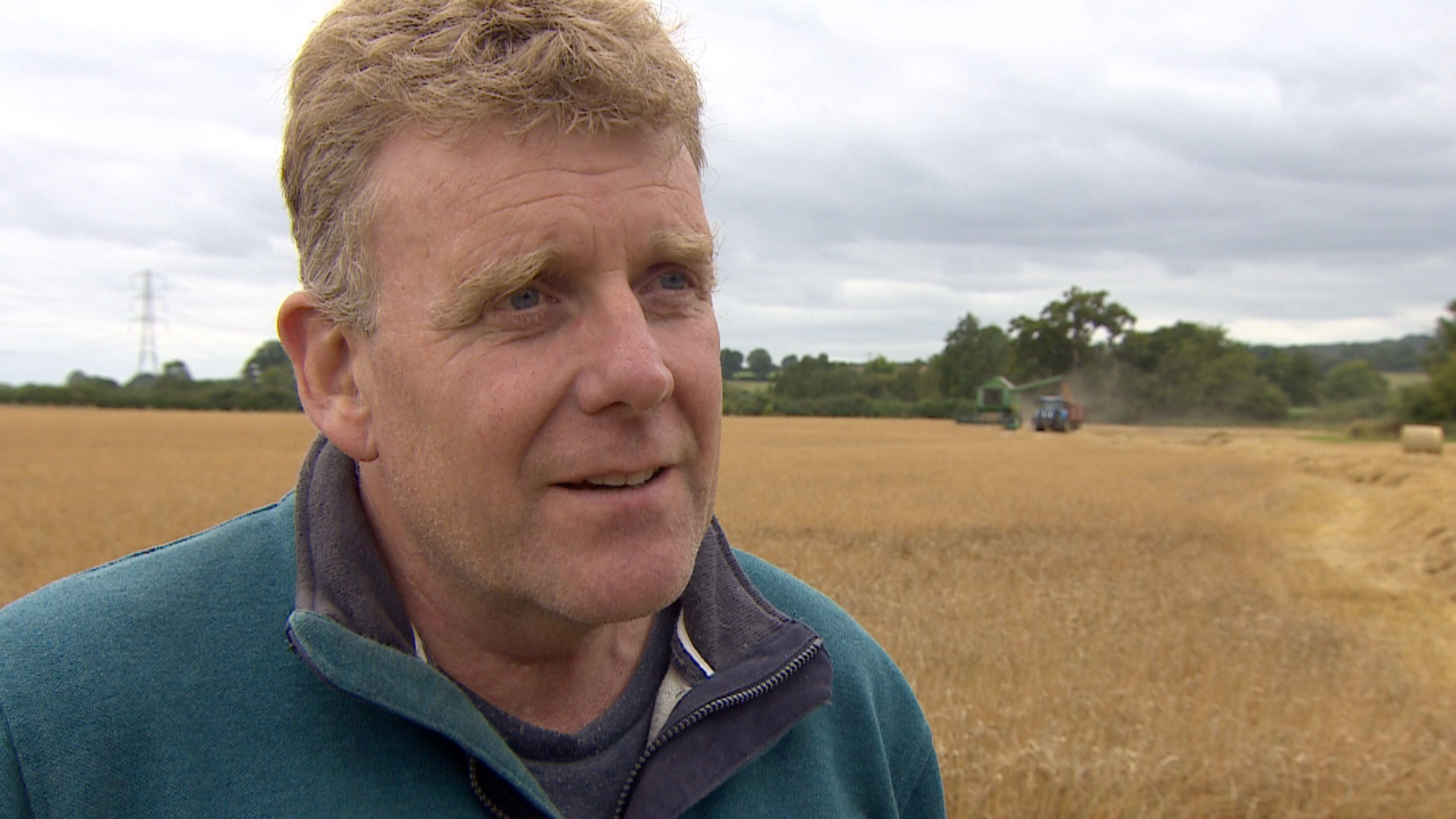 Farmer Patrick Twigger stands in a field of wheat, with a combine harvester working behind him