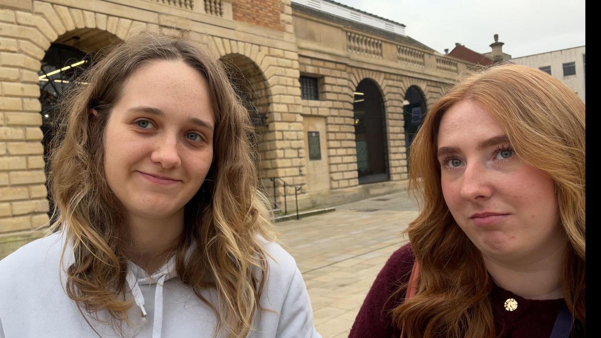 Two young women smile at the camera, one with long brown hair, the other with long ginger hair. The stone building which houses the Cornhill Market is visible in the background.