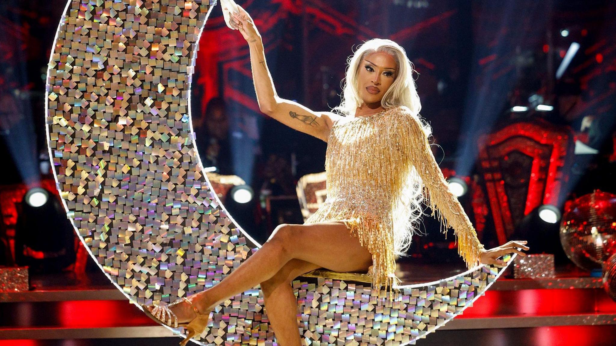 Tayce on stage on Strictly sitting cross-legged on a glittering crescent moon-shaped seat in gold dress and gold heels