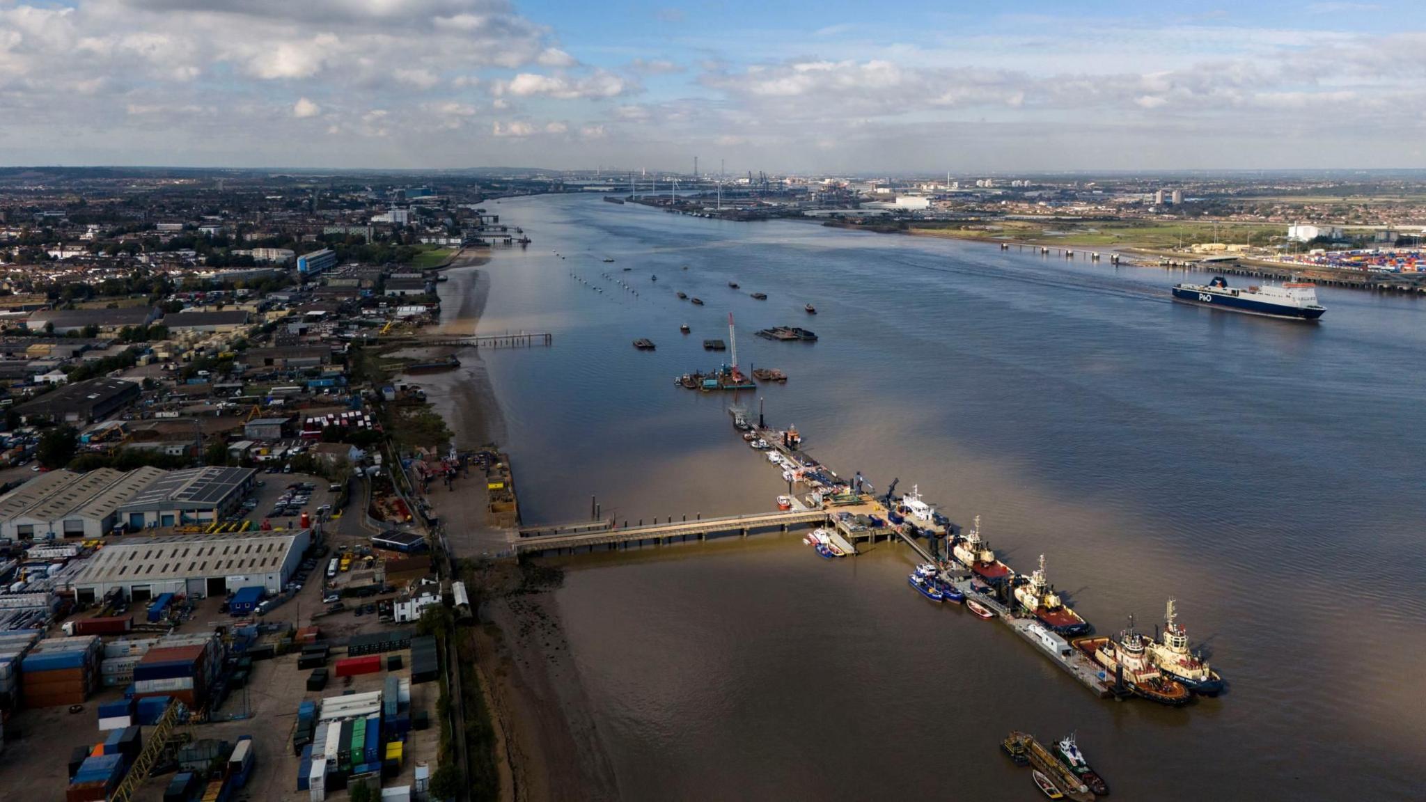 A view of the Thames with Gravesend on the left, with a pier and boats moored to is, with Essex and London visible on the right and horizon and a larger vessel sailing closer to the Essex bank of the estuary