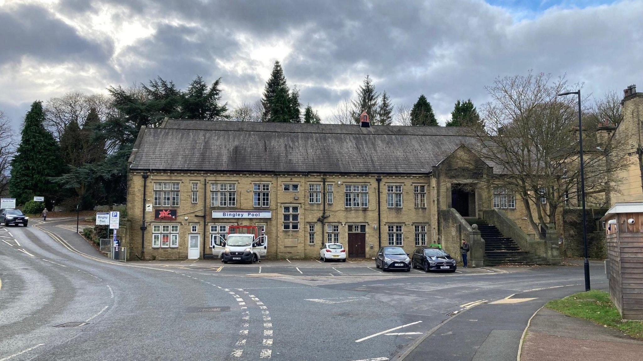 A two-story, Yorkshire stone building with a grey slate roof and trees at the back stands at the junction of two roads.