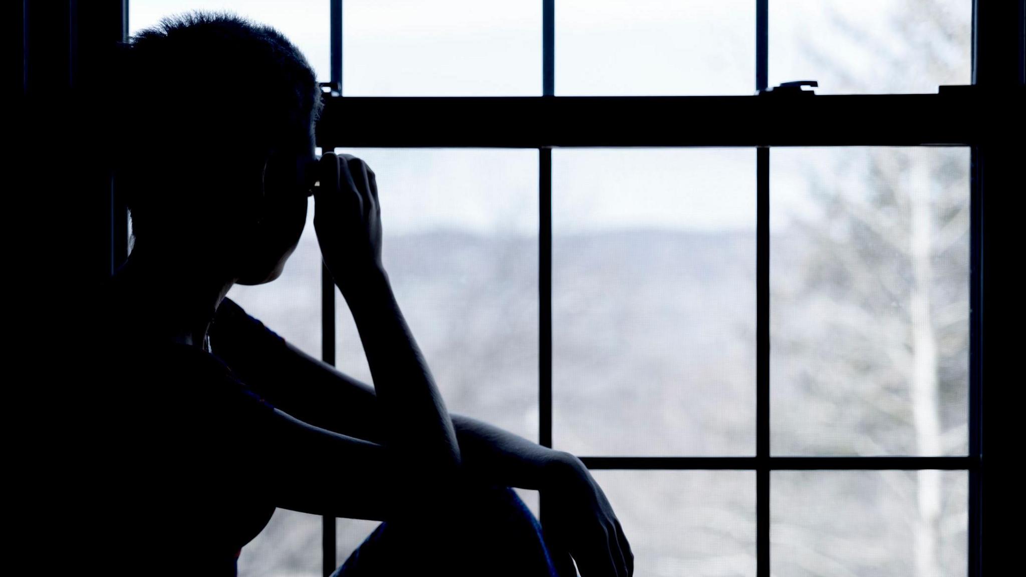 A shadow looks outside into the window. It is a black and white image. The young person has an arm to their head. 