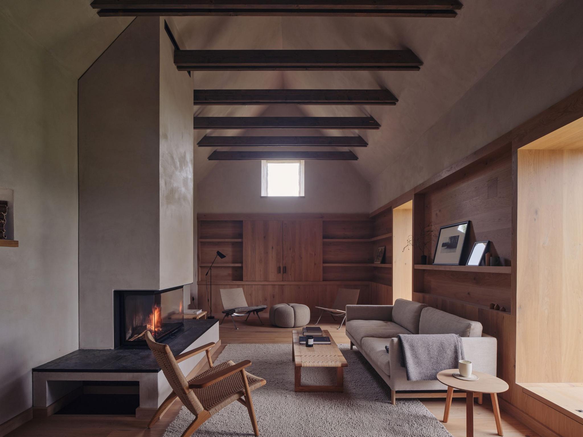 The living room has a blazing fire in a modern sleek fireplace, in front of a pale sofa. There are exposed oak beams overhead and pale wooden furniture