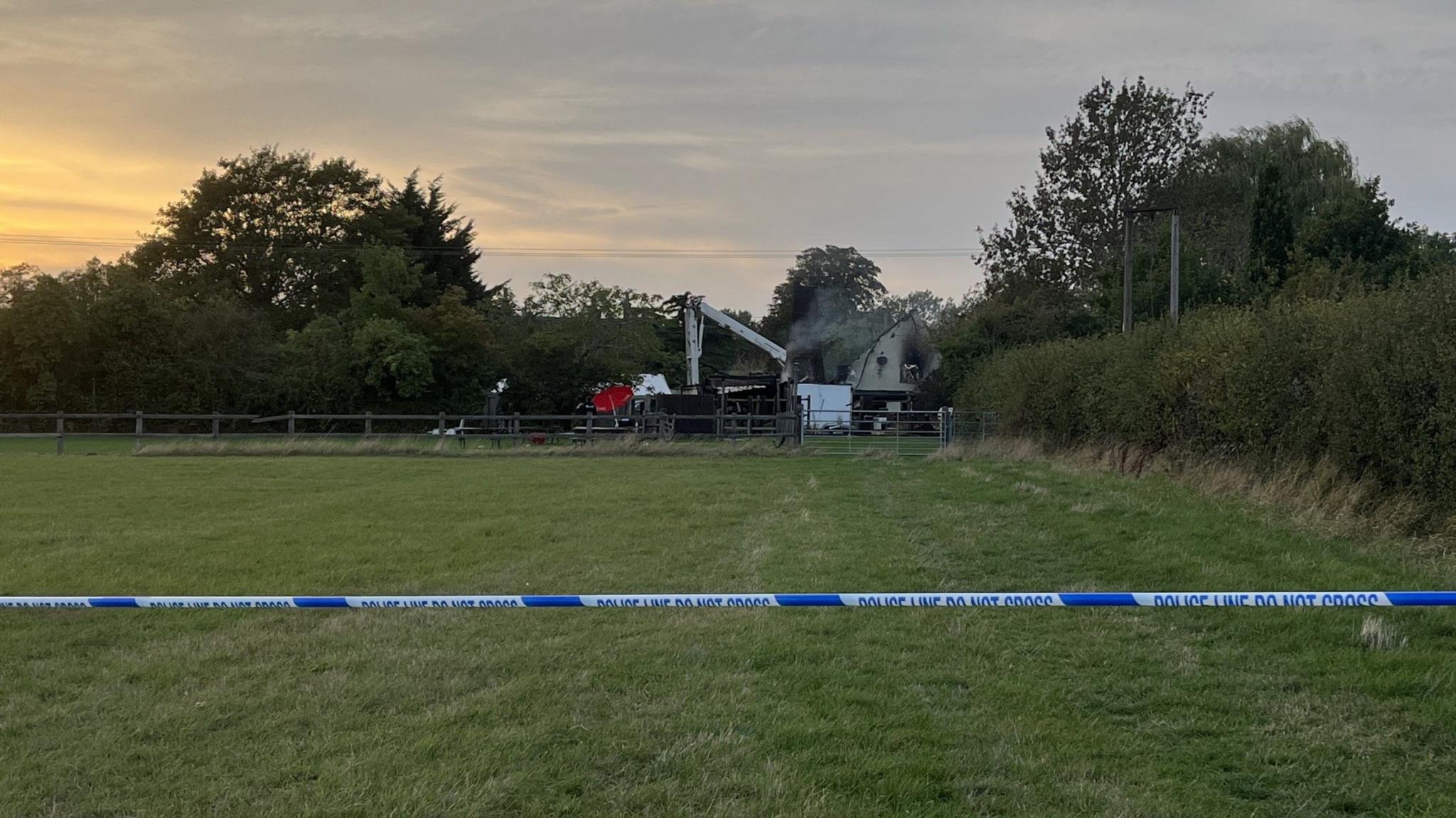 A picture from a field with blue and white police tap running across part of it to keep people out. Smoke can be seen billowing from a building in the distance. 