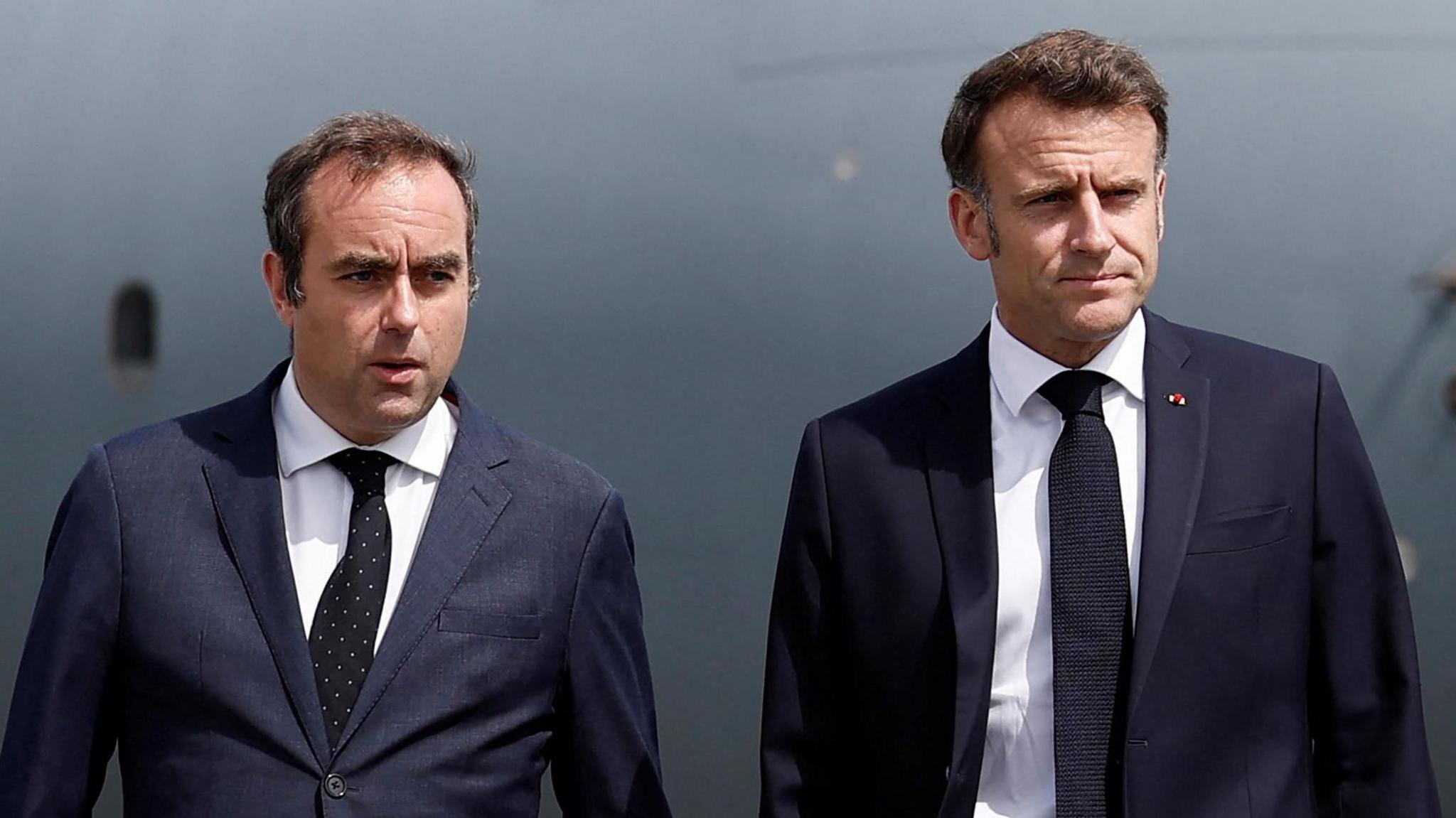 Sébastien Lecornu, who has dark hair and is wearing a dark suit, looks off to the centre while standing next to French President Emmanuel Macron, who is also wearing a dark suit.