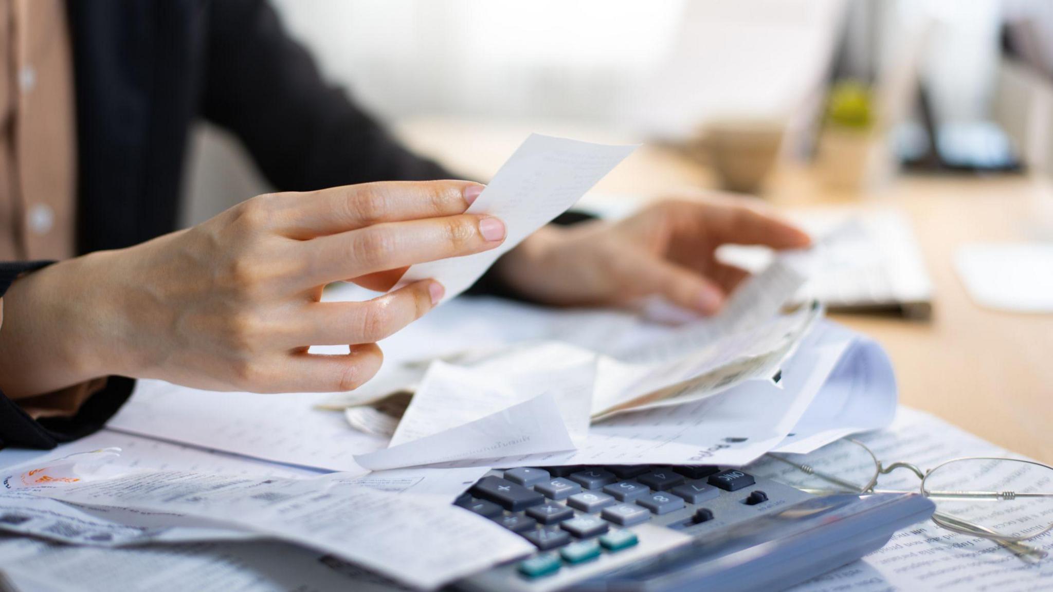 A person sits at a table wearing a dark coloured jacket and a light brown button up top. Only their hands are in focus. On the table in front of them is several sheets of paper and receipts, as well as a calculator and glasses.