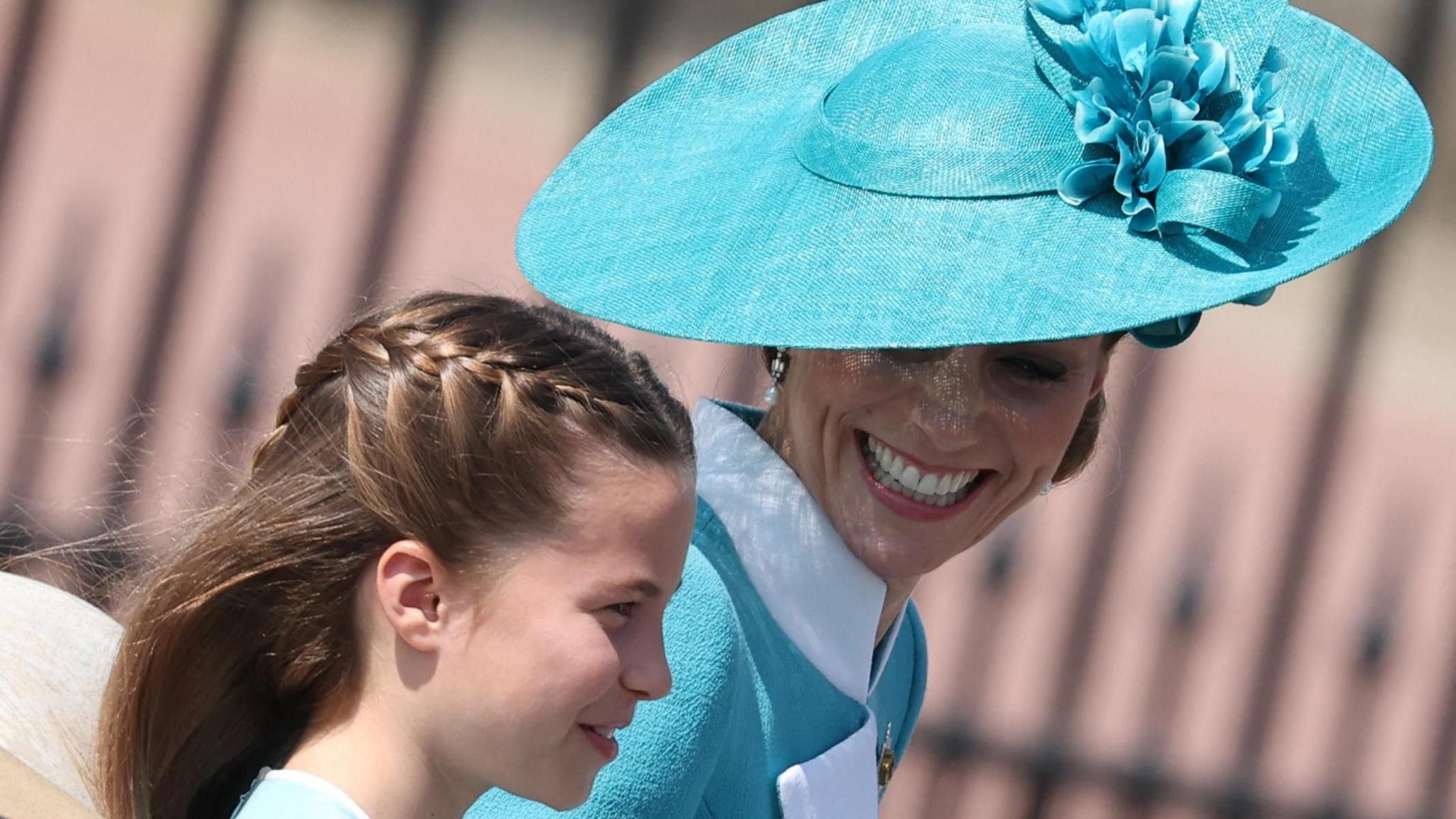 Princess Charlotte, who has long brown hair tied in a plait, smiles alongside her mother, the Princess of Wales, who is wearing a turquoise hat and dress