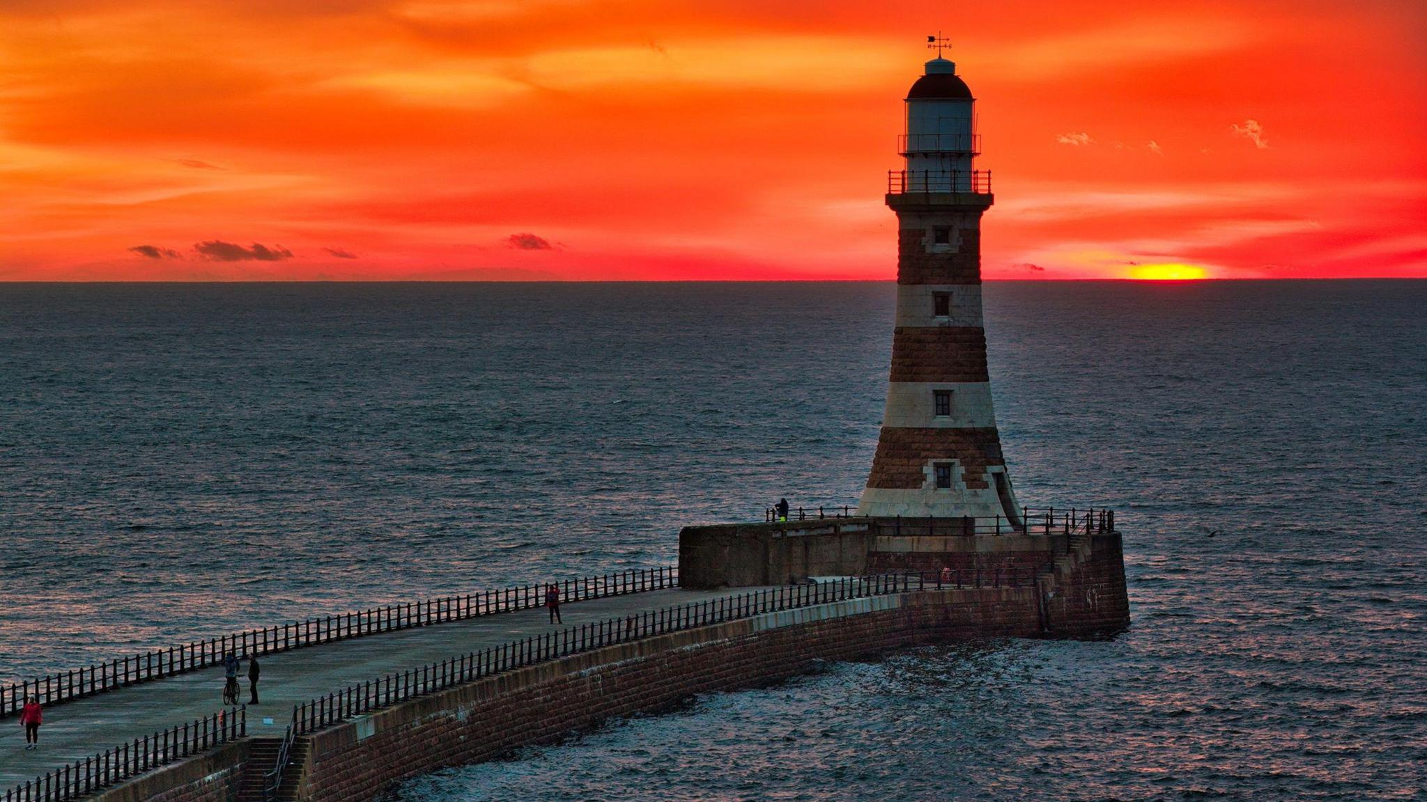 Roker Pier which is a long stone walkway into the sea. The lighthouse stands at the end of it with white and brown-looking stripes and is in shadow. There are people walking across the pier. The sea is calm around it but the sky is a bright red and orange colour.