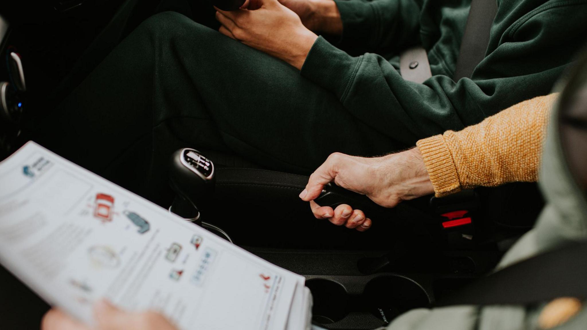 Two people sit side by side in the front of a car. The image is cropped tightly in on their legs and lower body. The passenger is holding paperwork which has diagrams for driving manouvres on it. They are pulling the handbrake up. The driver has both hands on the bottom of the steering wheel.