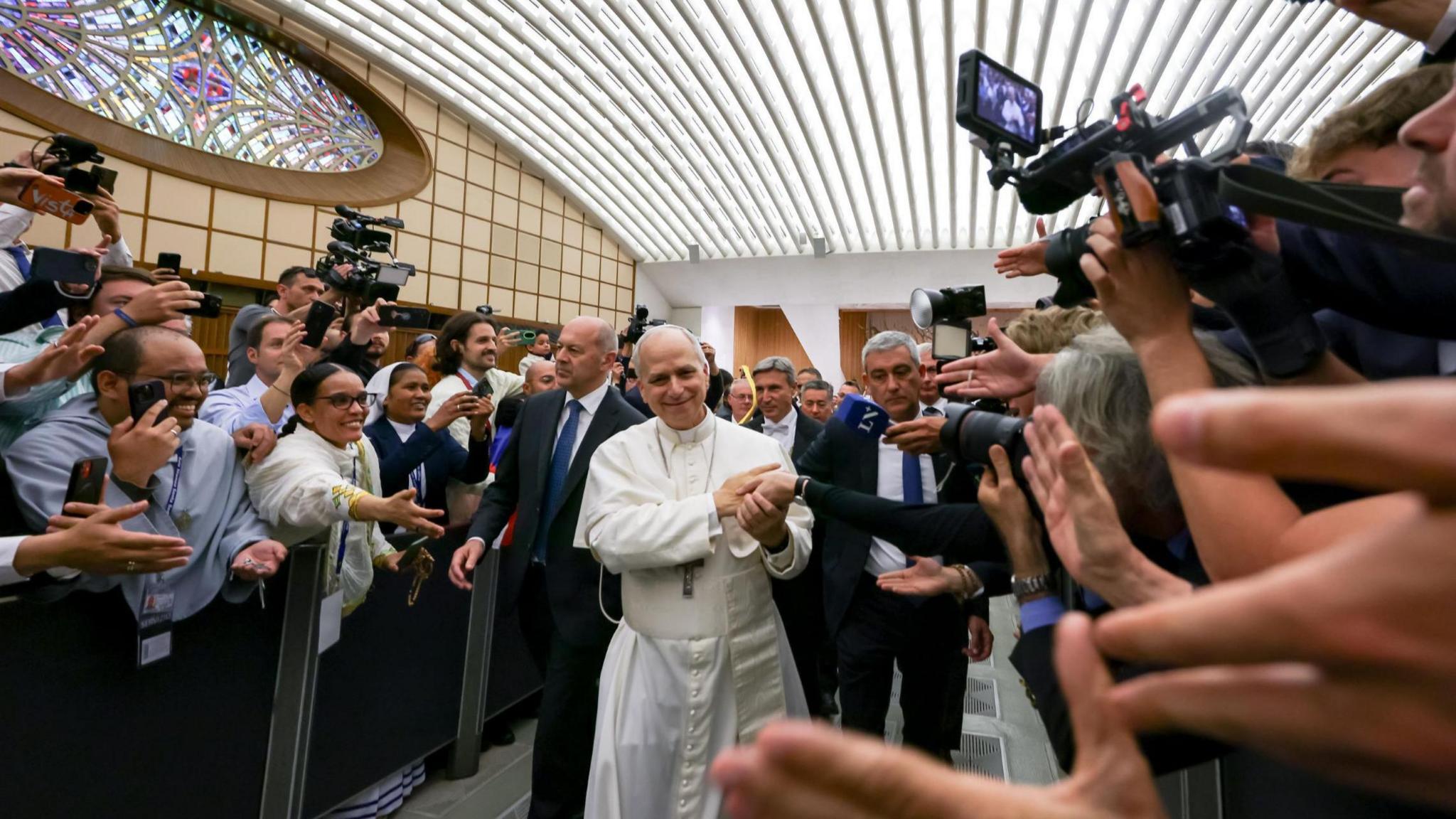 In the Nervi Hall at the Vatican, Vatican City, the pope in white walks past people who are behind a barrier, with their hands reached out towards him.