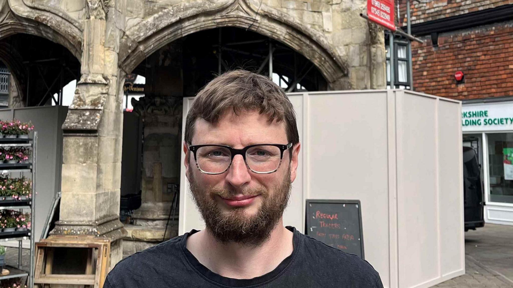 Sam Charleston standing in front of the Poultry Cross monument. He is wearing a dark T-shirt and has brown hair and glasses. Hoarding has been placed around the stone monument.