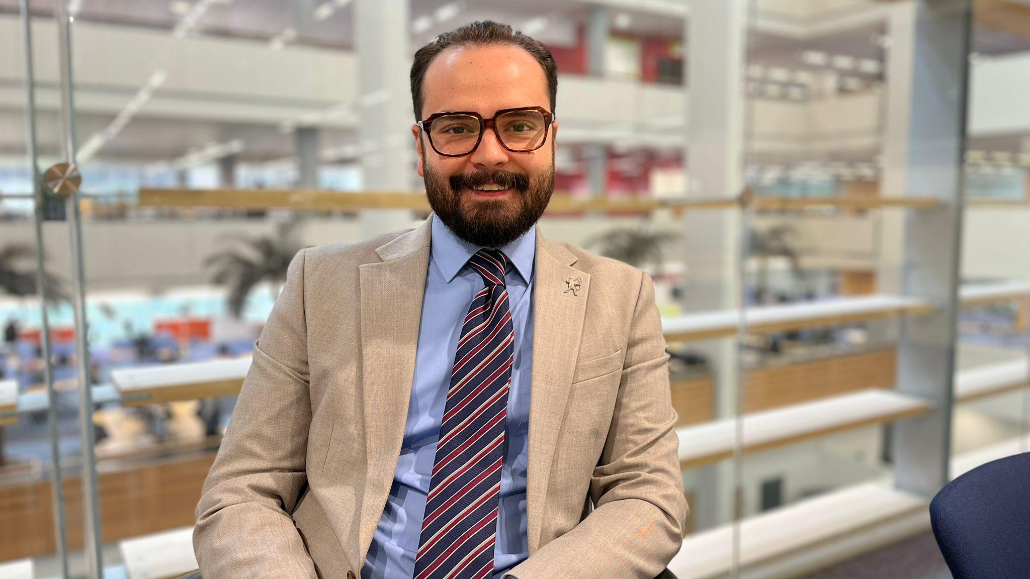 A smartly dressed man in a cream suit jacket, stropped blue, white and red tie and a sky blue shirt, sat down smiling for the camera in front of a wall of glass with desks below.