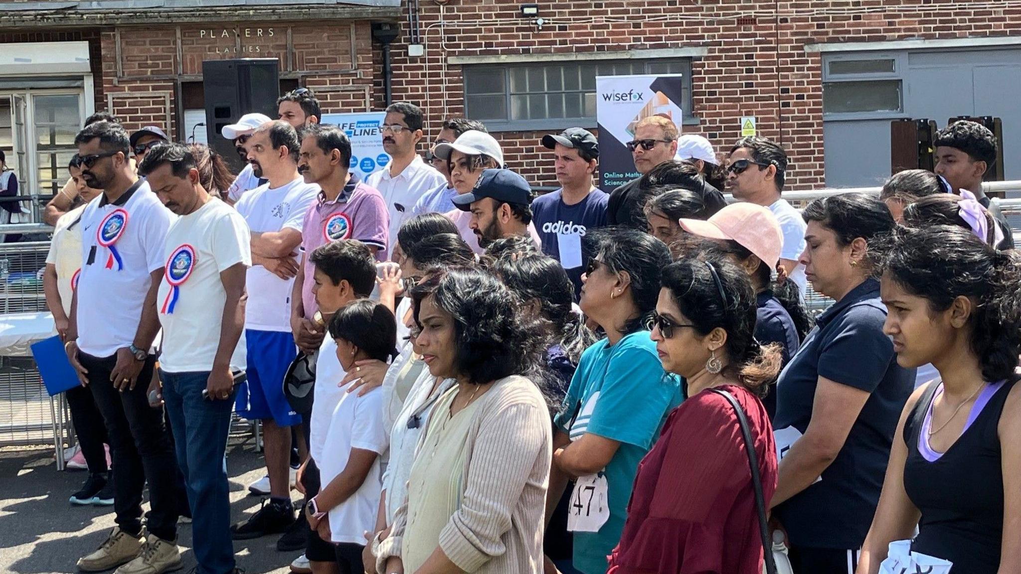 A crowd of people, some wearing sportswear, standing and looking solemn. There's a brick building behind them and it's a sunny day.