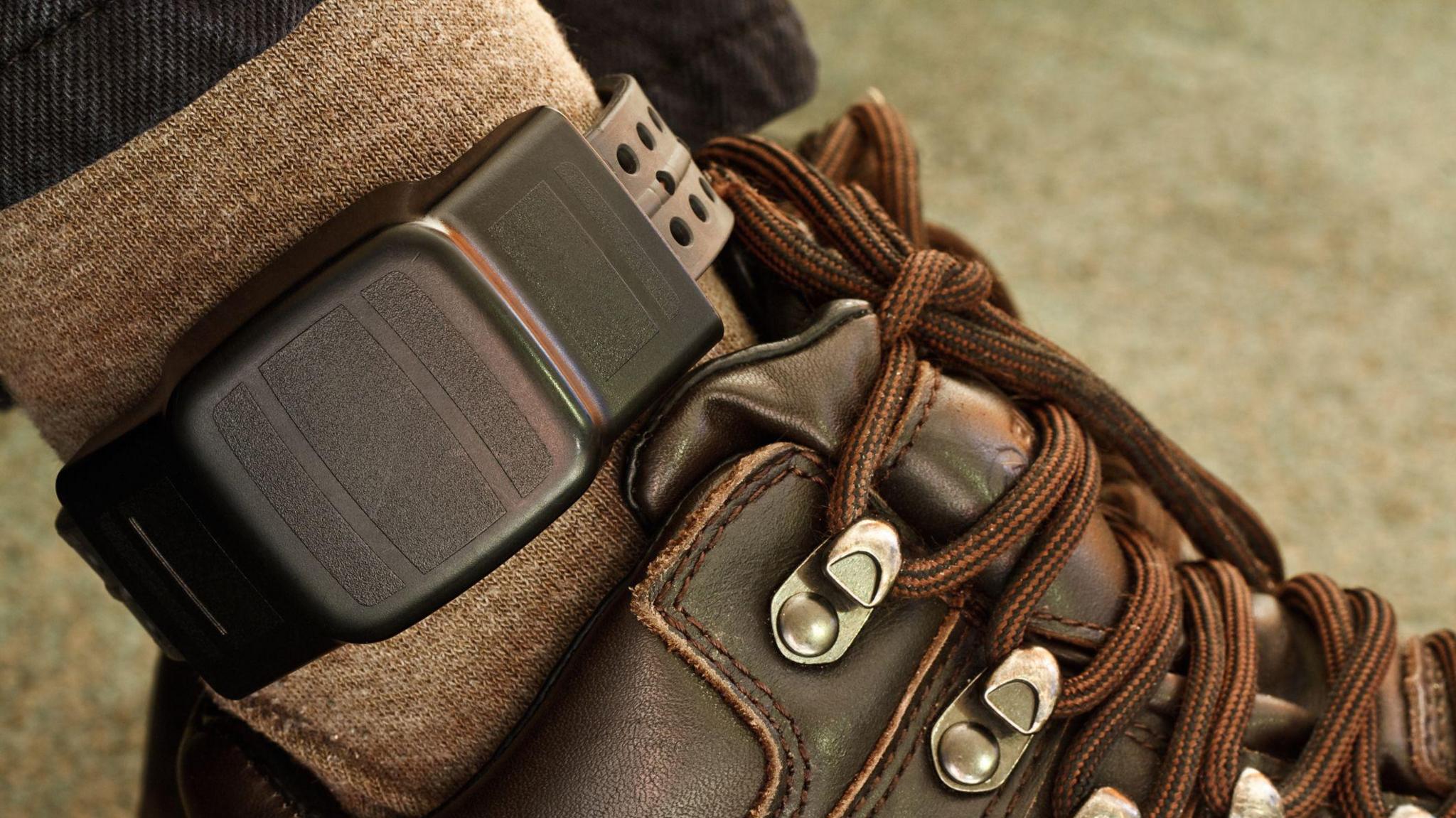 File image showing a black electronic GPS tag fitted around a person's ankle. Their brown socks and brown lace-up boots are visible