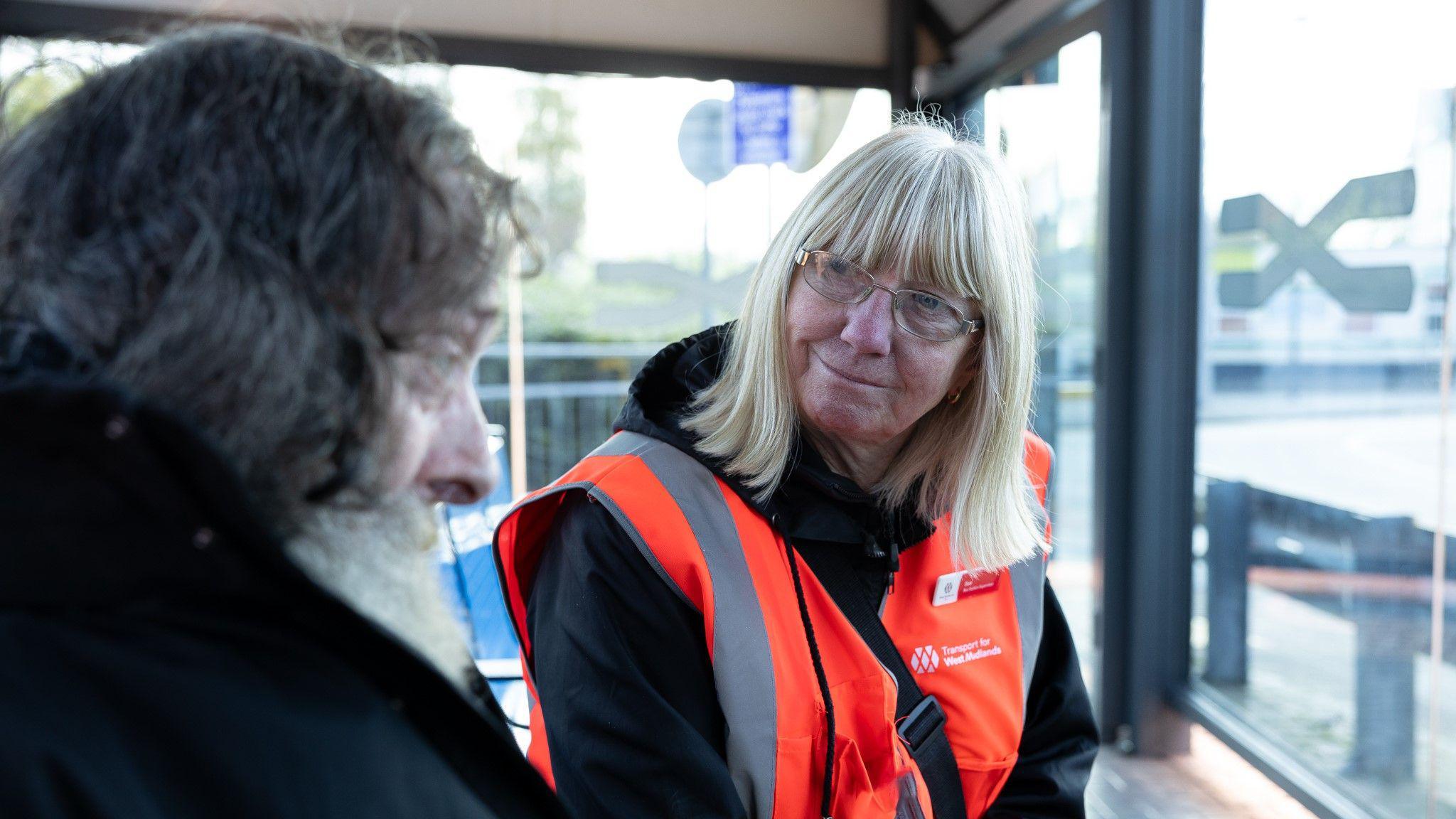 A lady with blonde hair and wearing glasses, looking at an older man with grey hair and a beard. They are sitting on a bench in a bus shelter