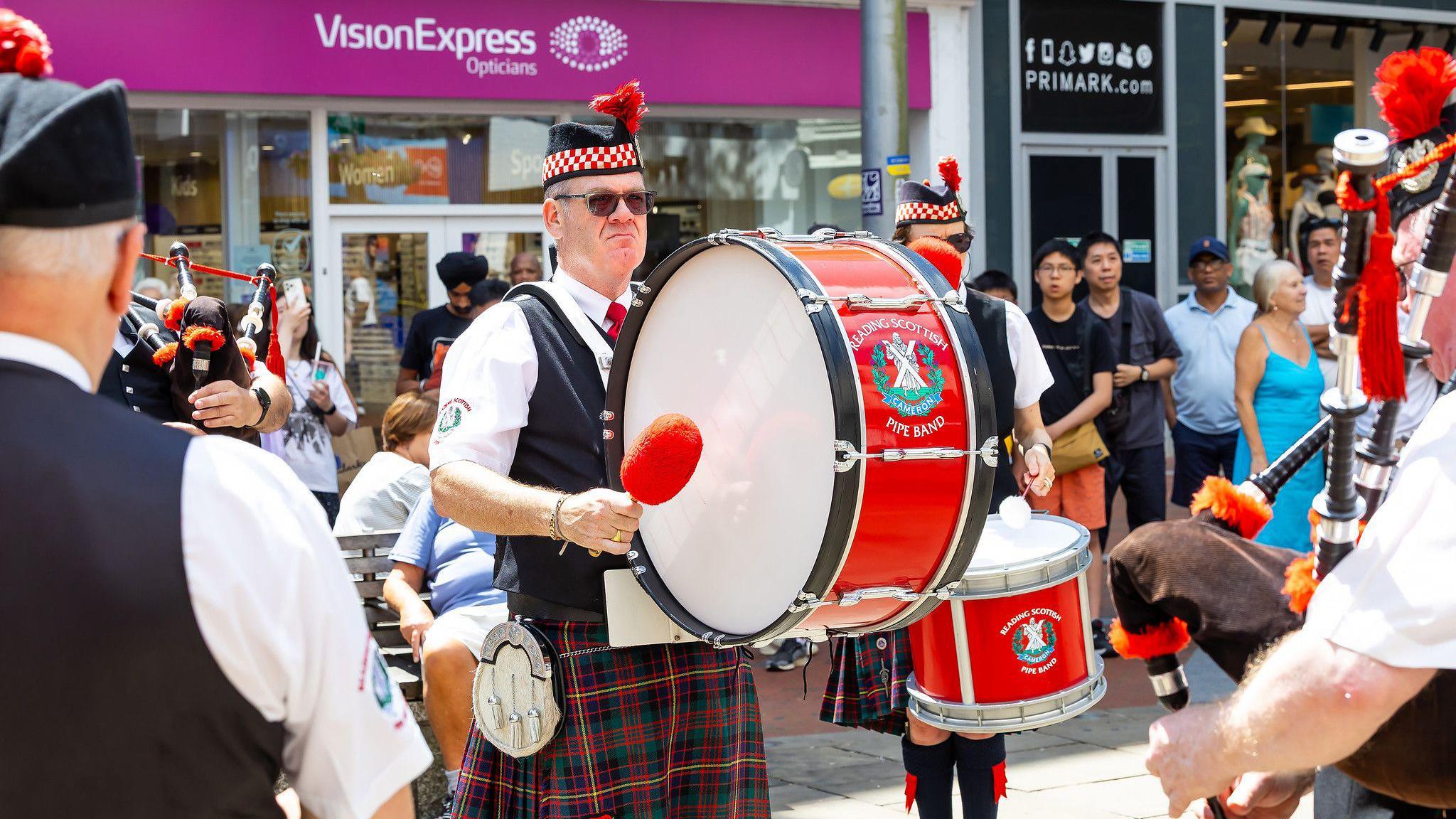 A man in traditional tartan clothing and kilt bangs on a large drums.