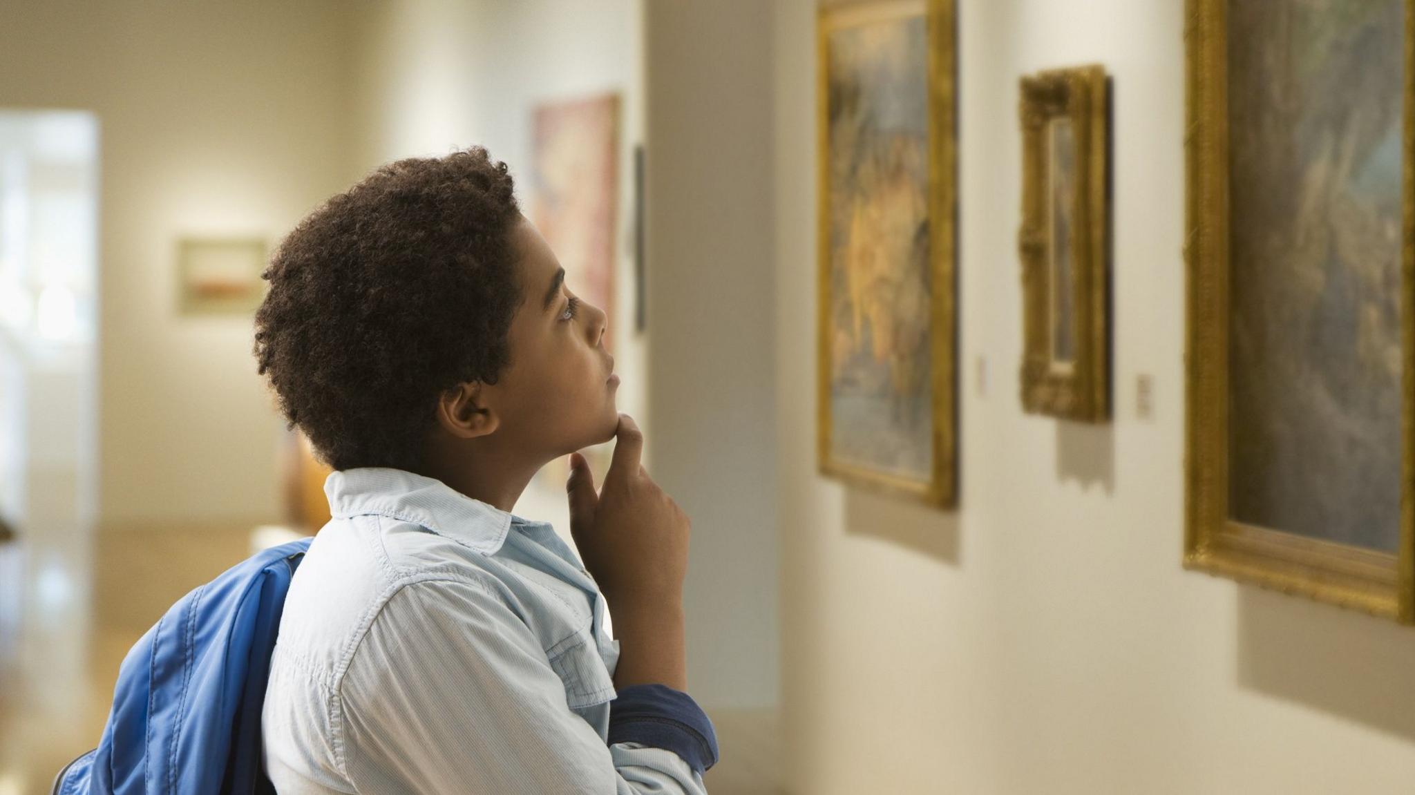 A boy rests his hand on his chin as he look at some paintings on display at an art gallery.