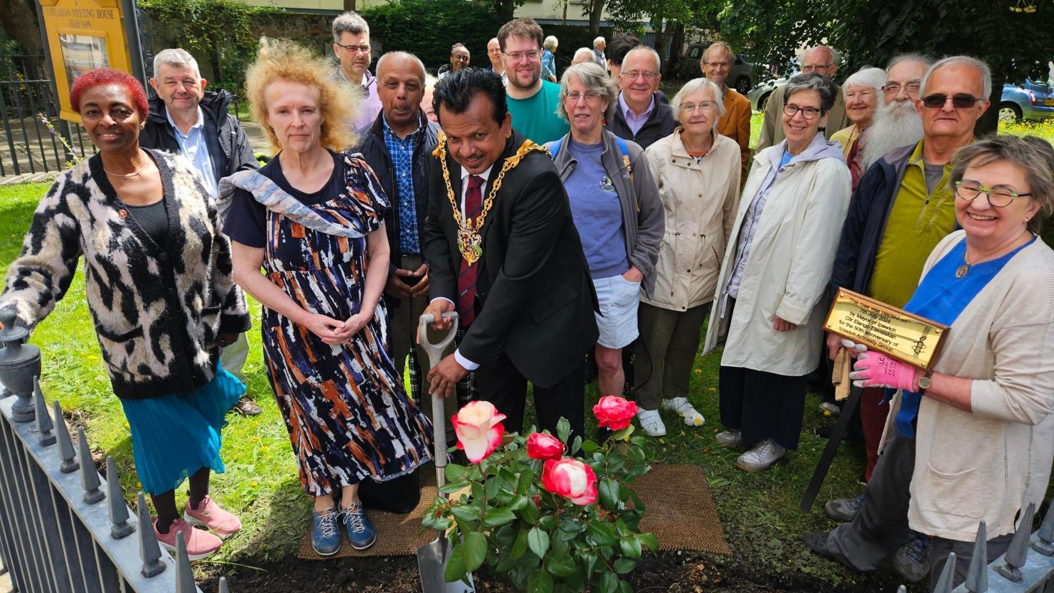 Ipswich Mayor Elango Elavalakan holds a spade, helping plants roses in a garden with a crowd gathered round.