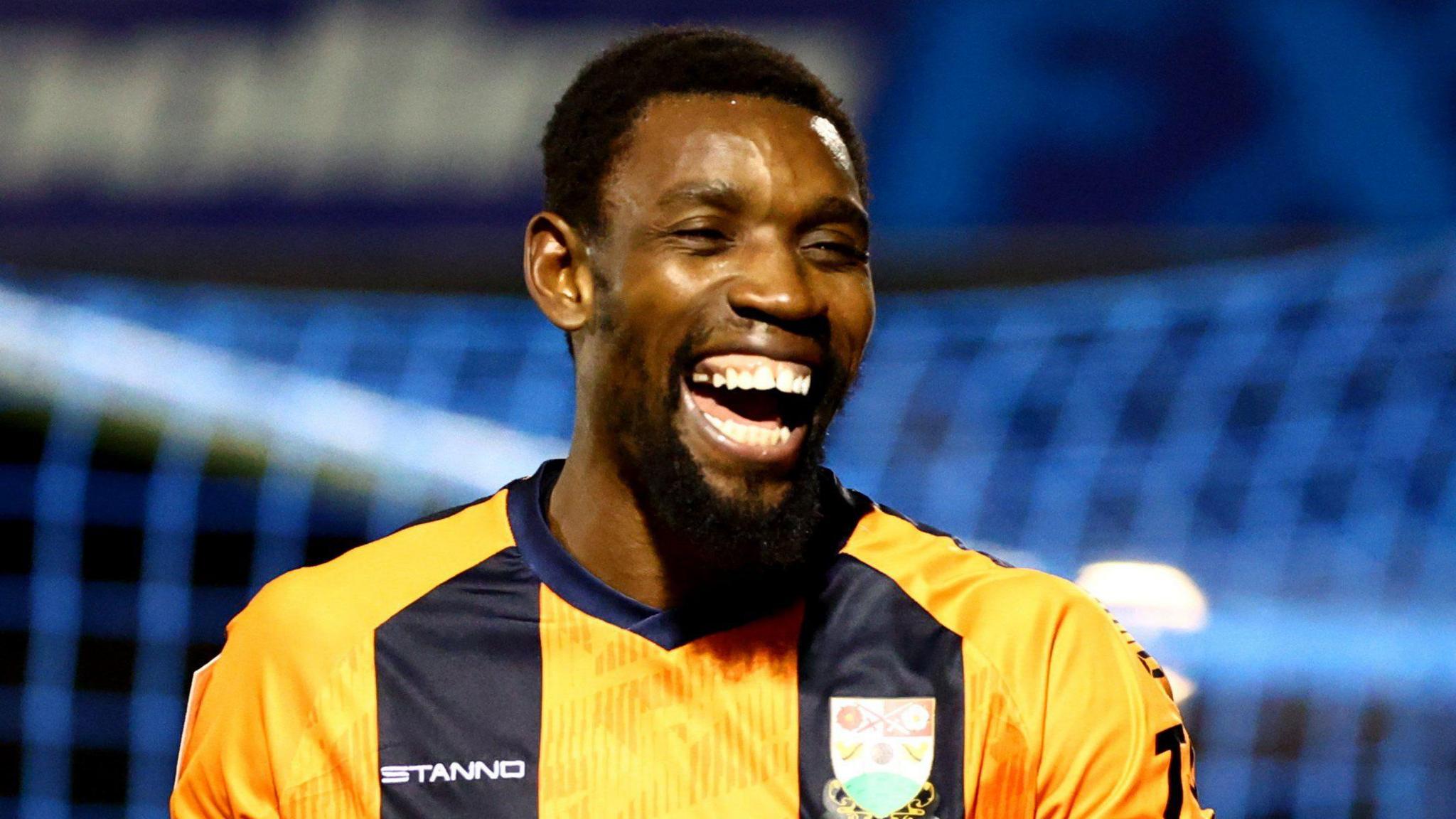 Lee Ndlovu with a big wide smile after scoring a goal for Barnet with a blue net in the background 