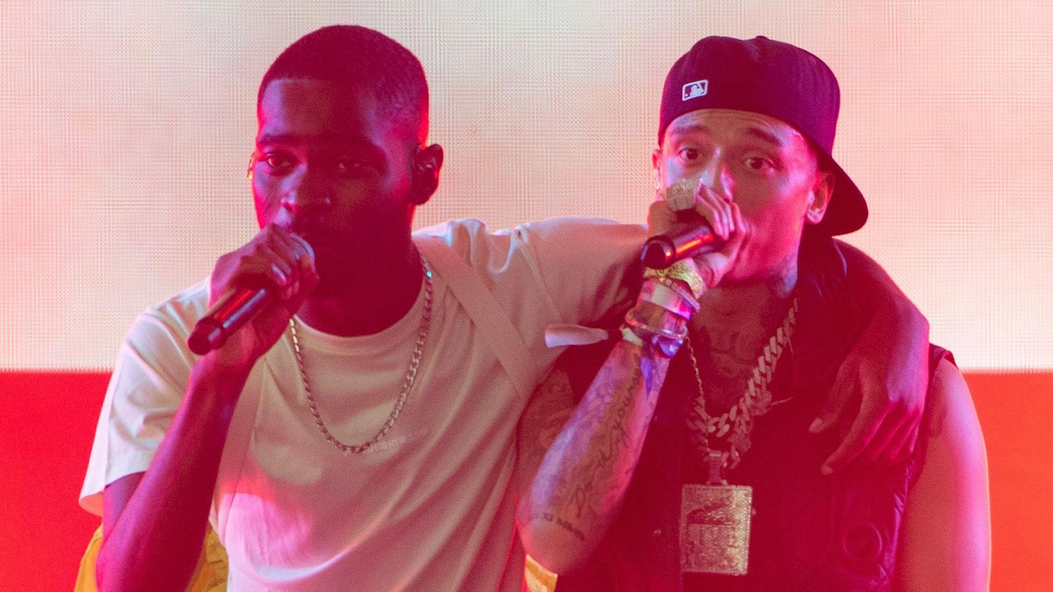 Central Cee on stage with Dave at Glastonbury in 2023. Dave, left, has his arm around Central Cee's shoulder as they both sing into microphones. Dave wears a white T-shirt and a pale pink backpack, while Central Cee has a black cap twisted backwards and wears a black gilet with a large chain necklace. 