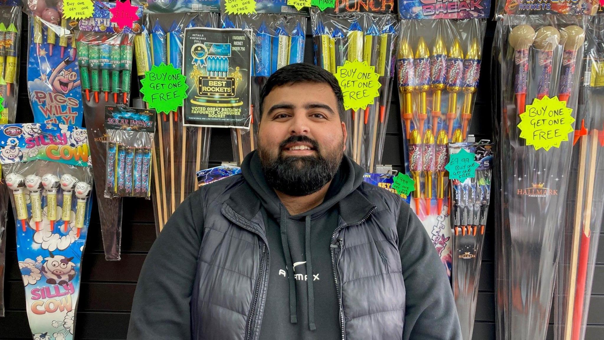 A man in a black bodywarmer and jumper standing in front of wall with colourful packets of fireworks on display.