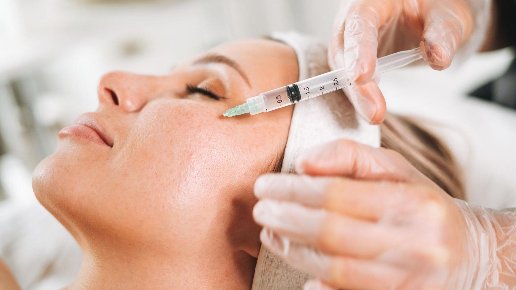 Gloved hands administering an injection with a syringe into the skin on a woman's face during a cosmetic or medical procedure.