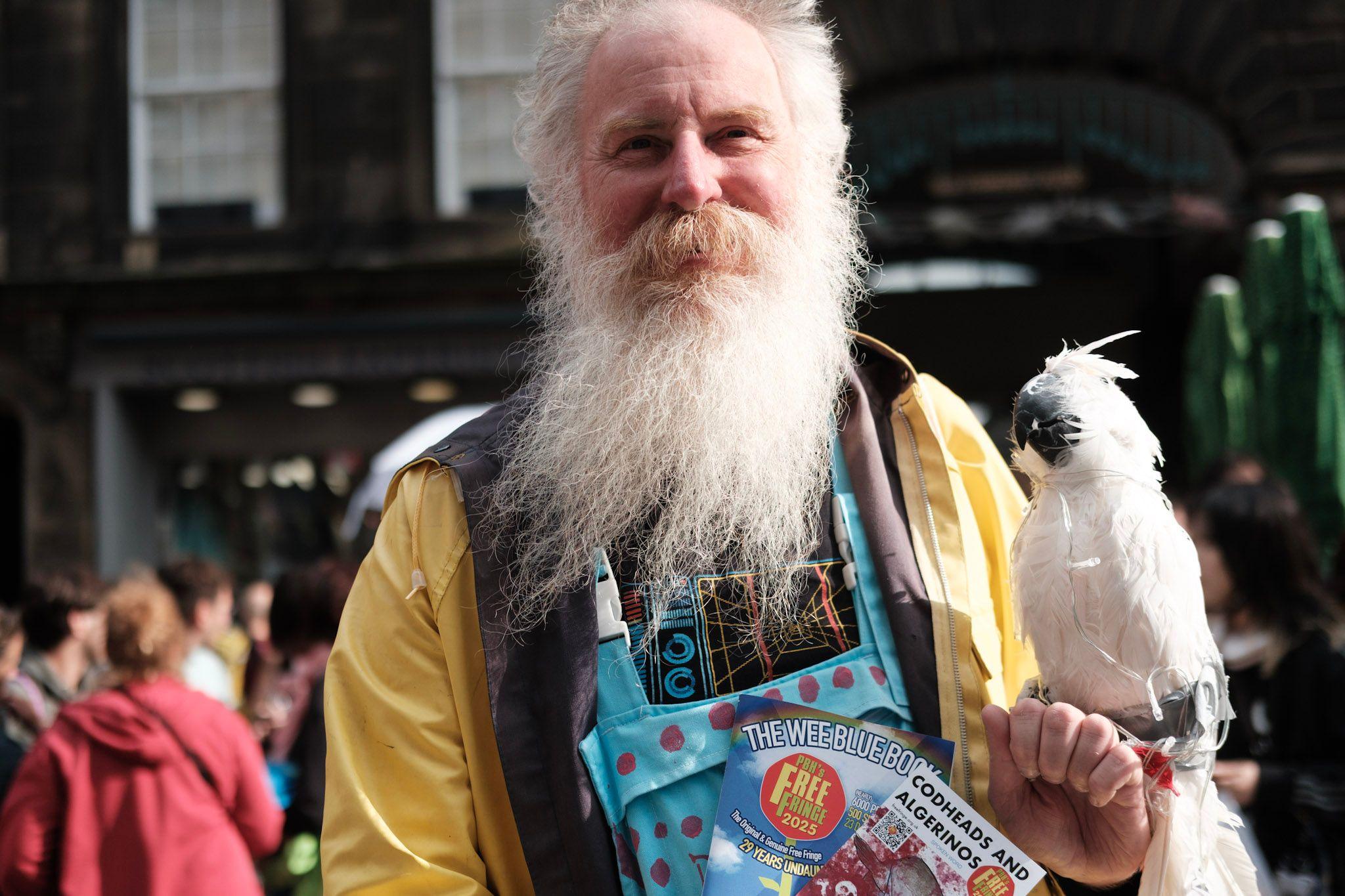 A man with a long, white beard holds a white cockatoo-type bird as he hands out flyers. He is wearing a yellow waterproof jacket and brightly coloured dungarees. He also has a blue and red spotty bag round his neck