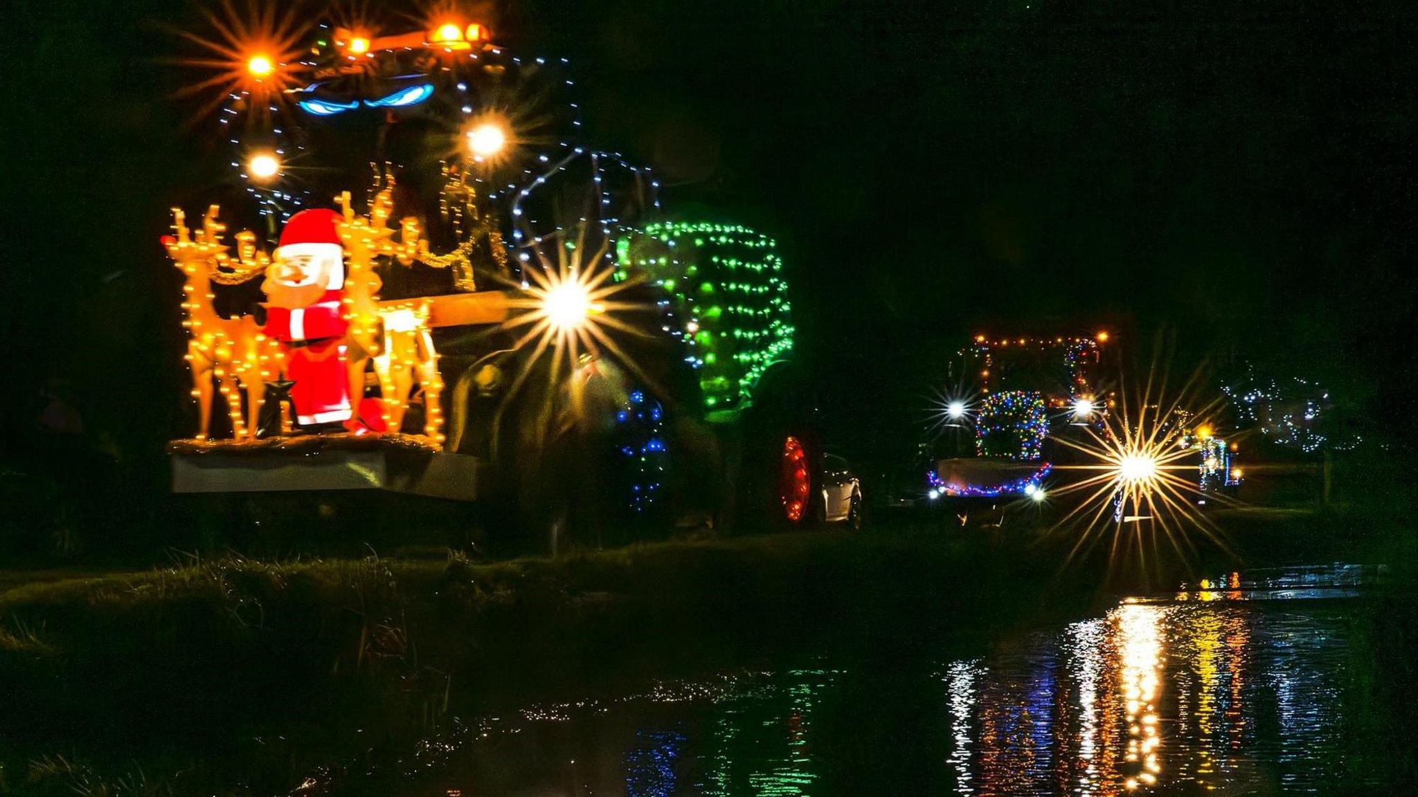 A procession of tractors driving at night. The closest one to the camera has a red Santa Claus and reindeer either side lit up at the front. 