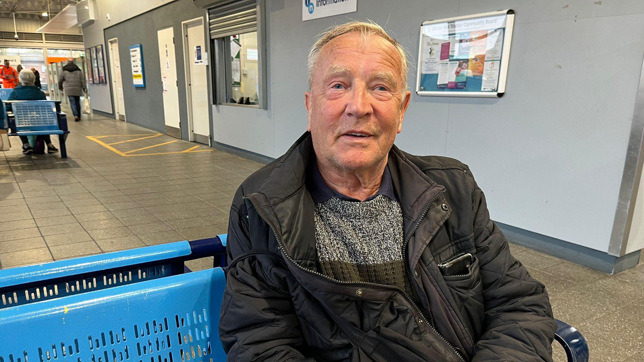 Brian Fisher sat at Bilston bus station. He is sat on a blue bench and wearing a black coat with a grey and green jumper. 