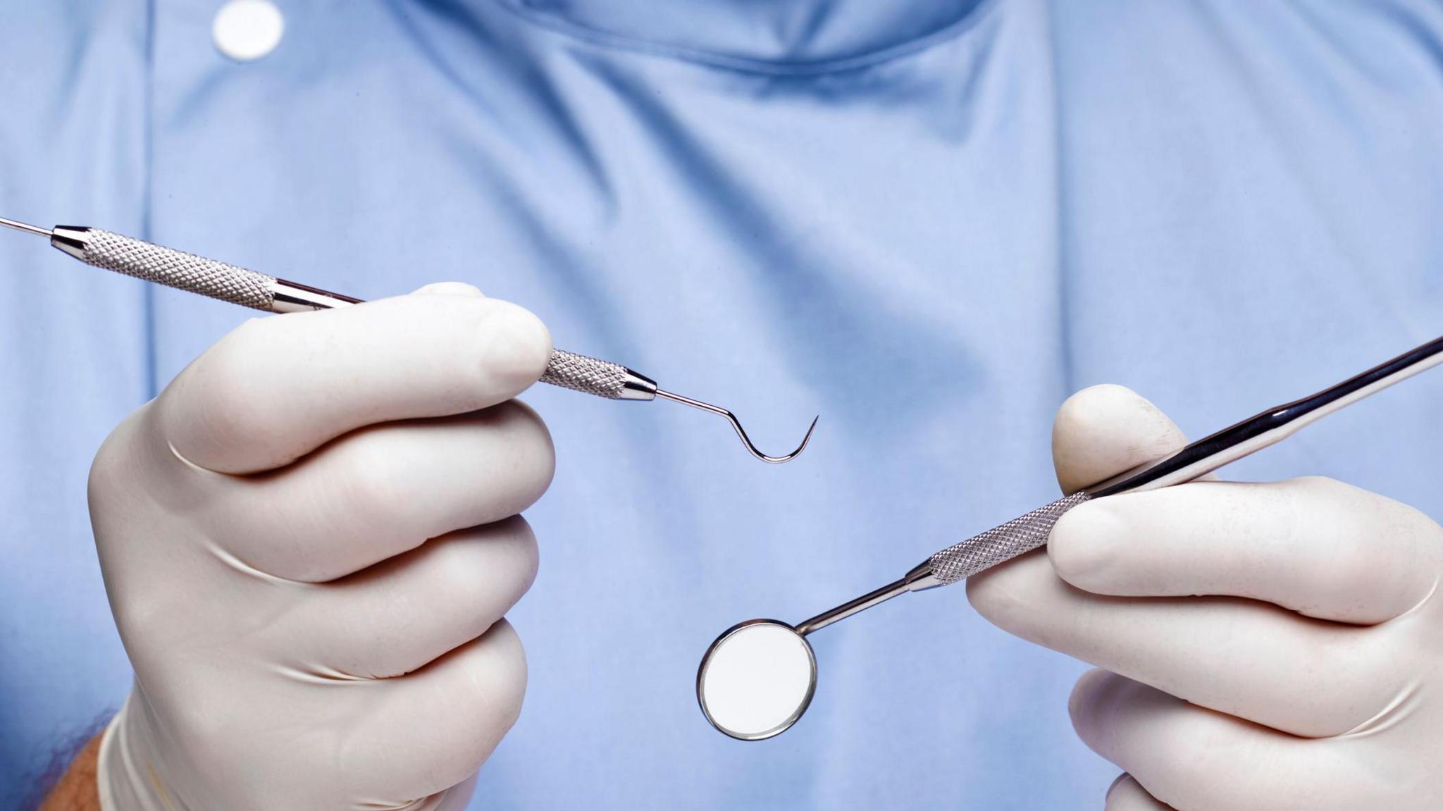 Close-up of a dentist wearing a blue gown and white latex gloves, holding dental implements including a mirror.