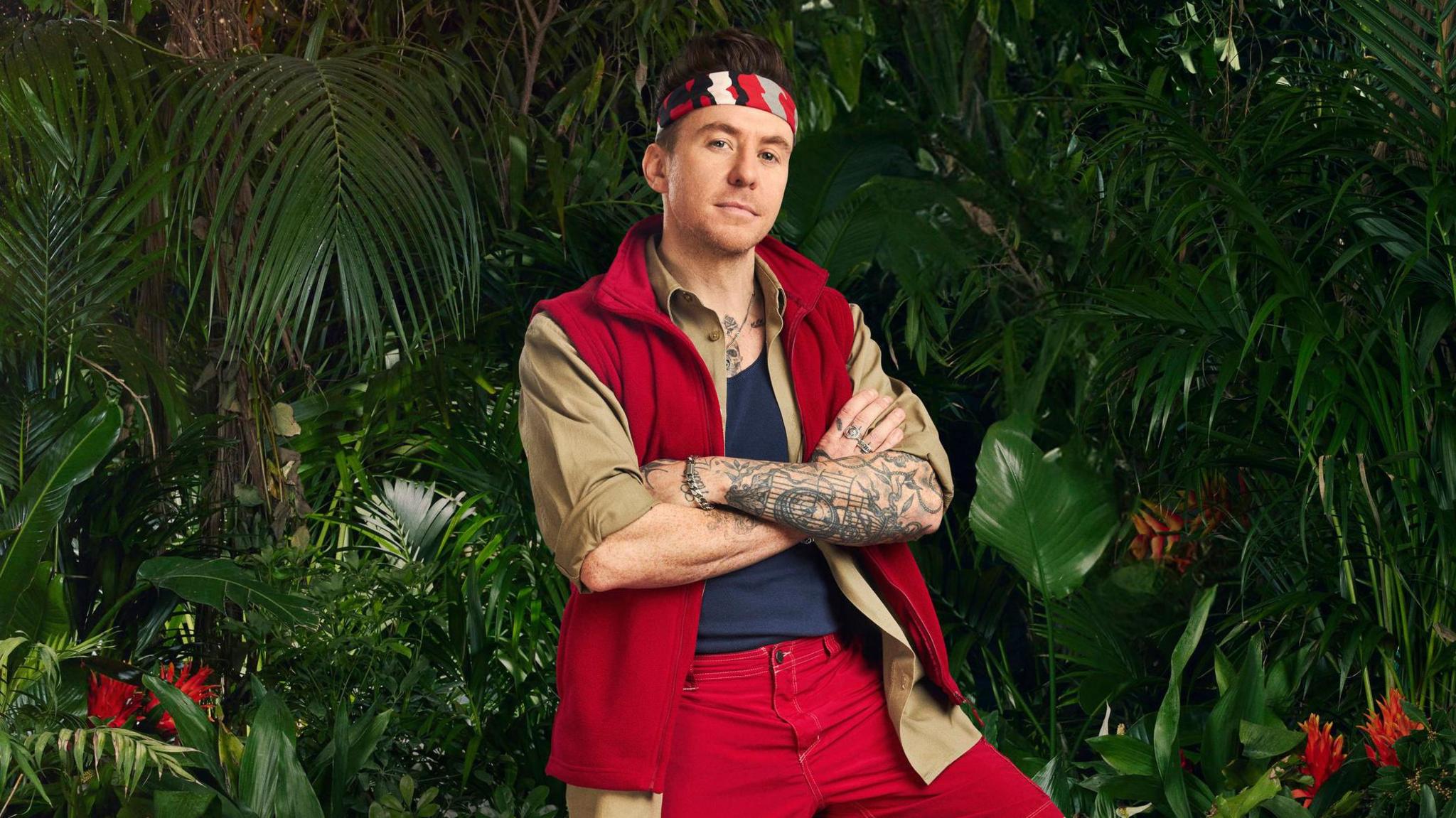 Danny Jones with his arms folded, wearing his jungle outfit and a bandana, surrounded by plants 