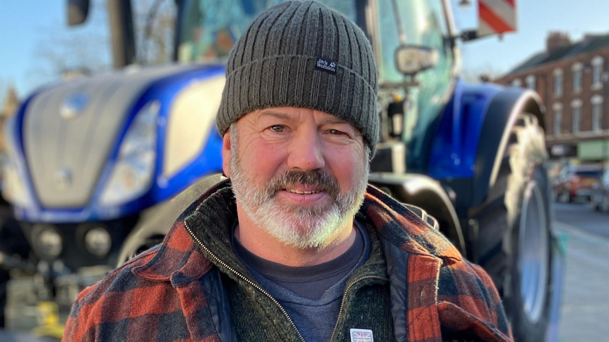 A man with a grey beard and wearing a woolly hat and red checked shirt stands in front of a blue tractor.