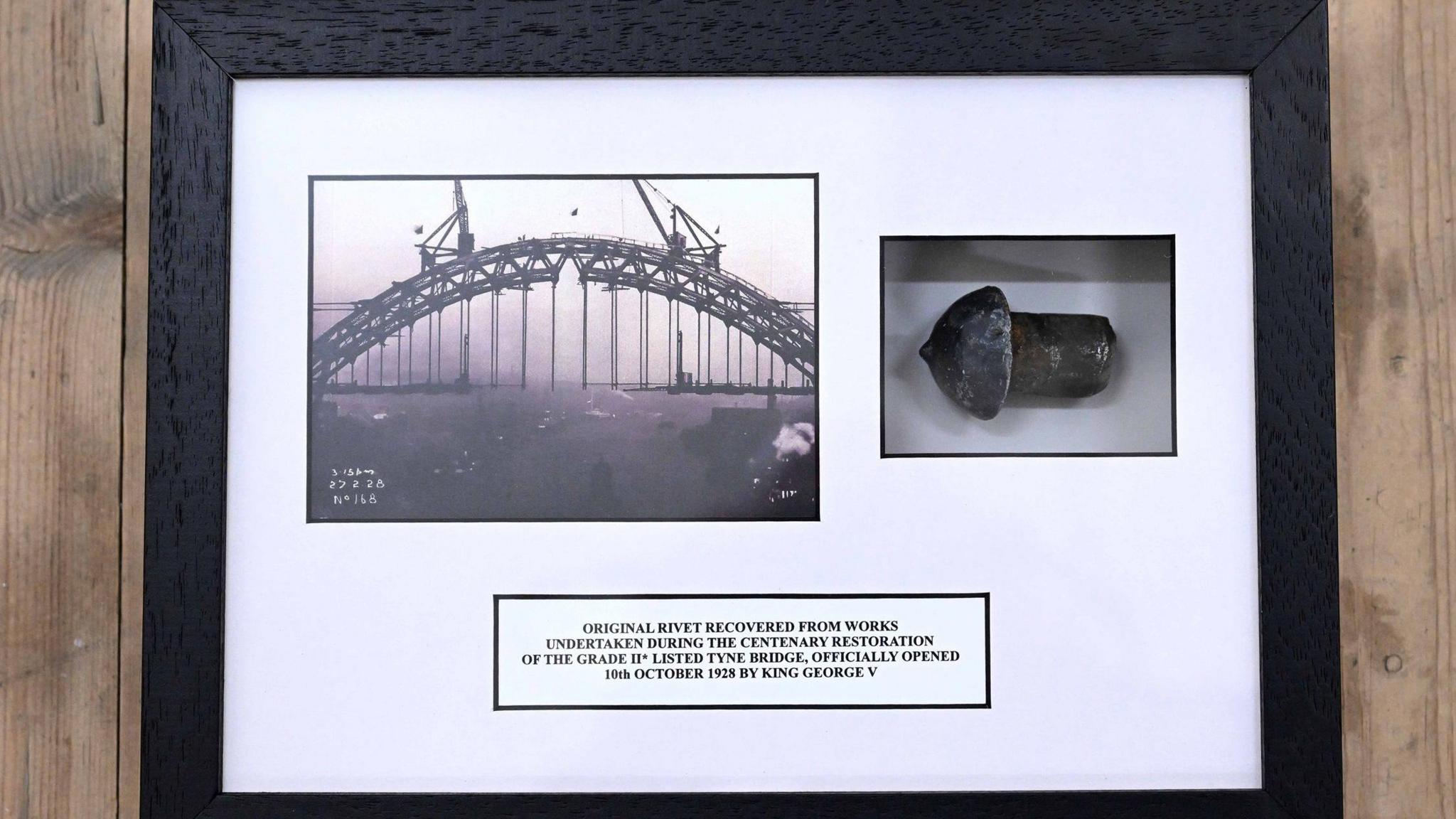 A framed picture of the Tyne Bridge being constructed in black and white. Next to it is the rivet which is a small piece of metal with a domed top. Written below is a plaque which reads: "Original rivet recovered from works undertaken during the centenary restoration of the Grade II* listed Tyne Bridge, originally opened on 10th October 1928 by King George V."