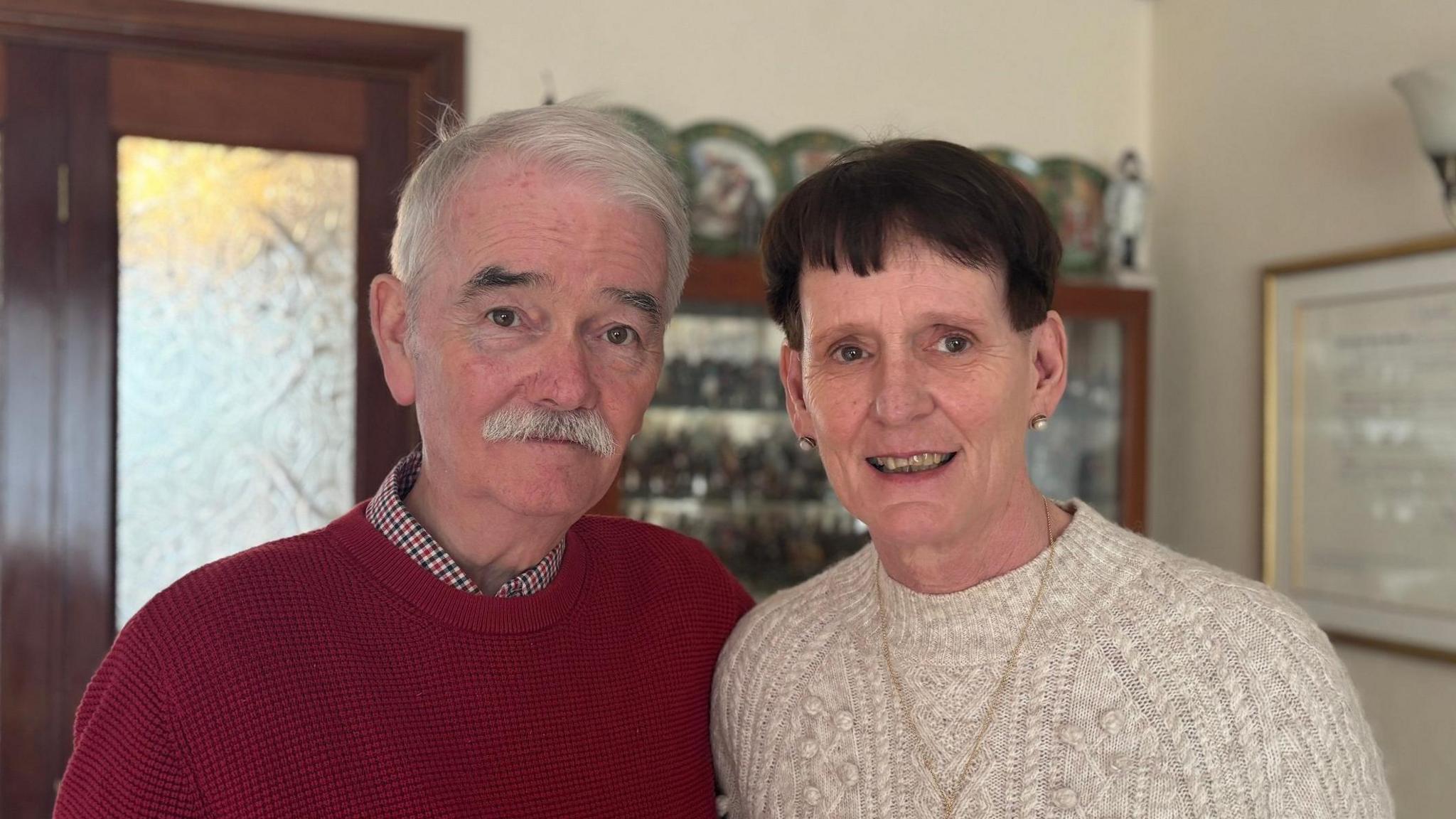 Peter Cunningham has grey hair and and grey moustache and is wearing a red round neck sweater. Patricia Cunningham has short dark hair. She is wearing a beige knitted round neck sweater and had pearl earrings.