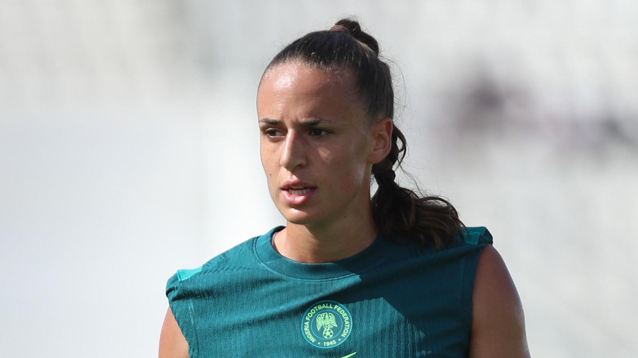 A head shot of Nigeria defender Ashleigh Plumptre shows the defender from the shoulders up during training. She is wearing a dark green training top with a Nigeria Football Federation crest in the centre of the chest and has the sleeves rolled up to reveal the tops of her shoulders. Her hair is pulled back in a ponytail which is falling across her left shoulder while there is a slight glisten of sweat on her forehead as the sun shines on it