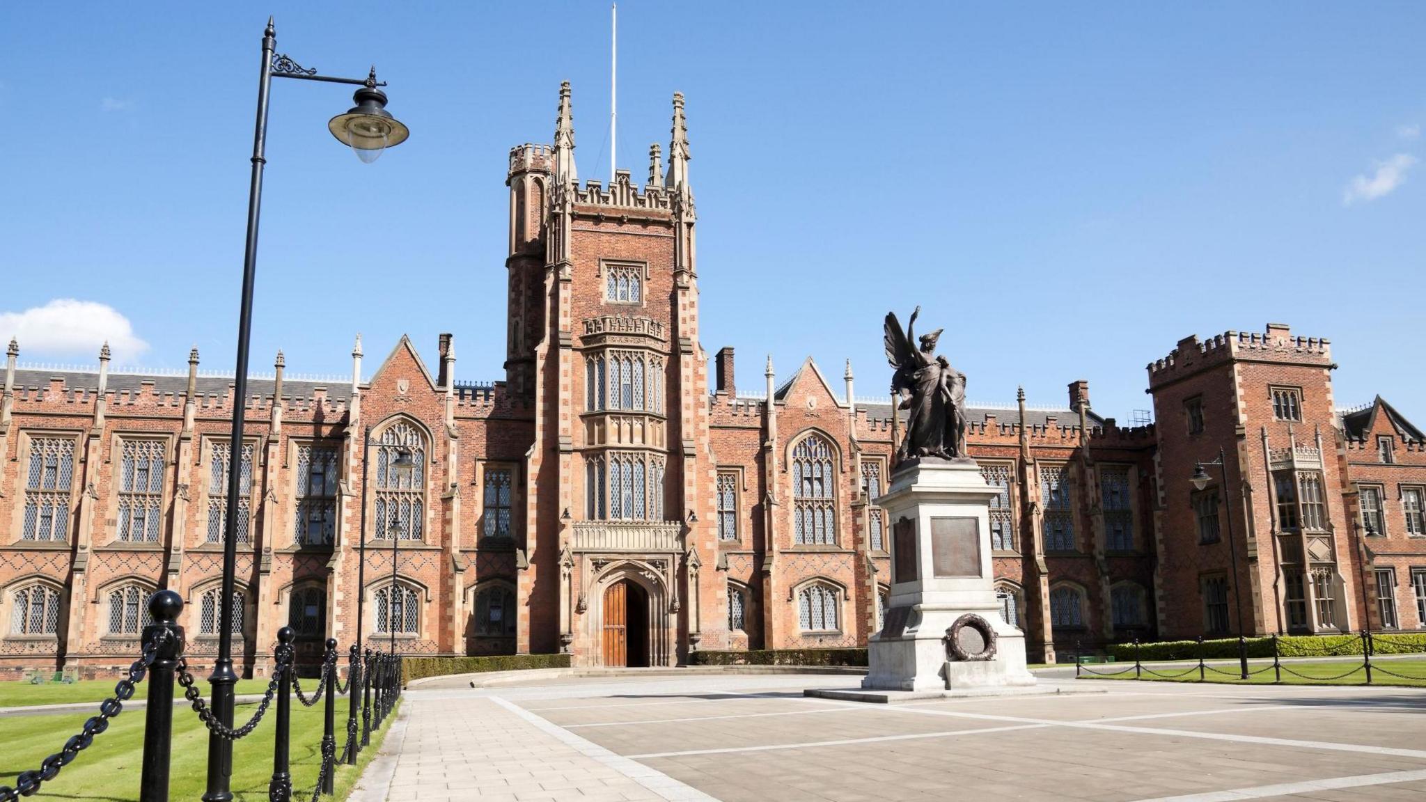 The exterior of Queen's University Belfast's Lanyon Building taken from it's front courtyard. It is a large, Gothic/Tudor style two-storey, red brick building with large panelled windows along it's exterior. There is a war memorial with a white stone plinth just in front of the building's large wooden double doors. It is a sunny day, with minimal clouds and mowed grass in front of the building.