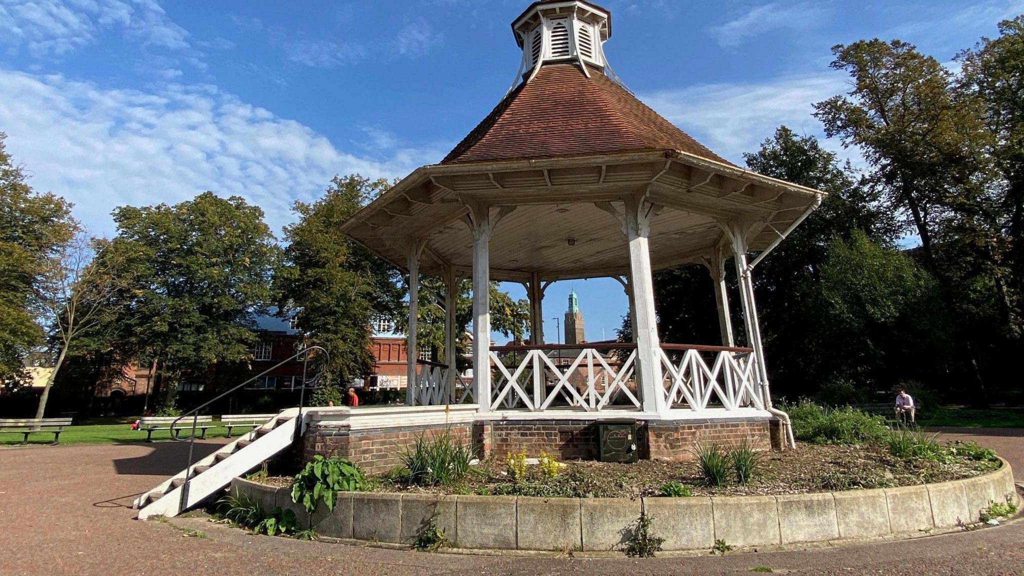 A Bandstand on top of a stone base. The wooden structure has been painted white and has steps up to it. Around it are trees and benches. 